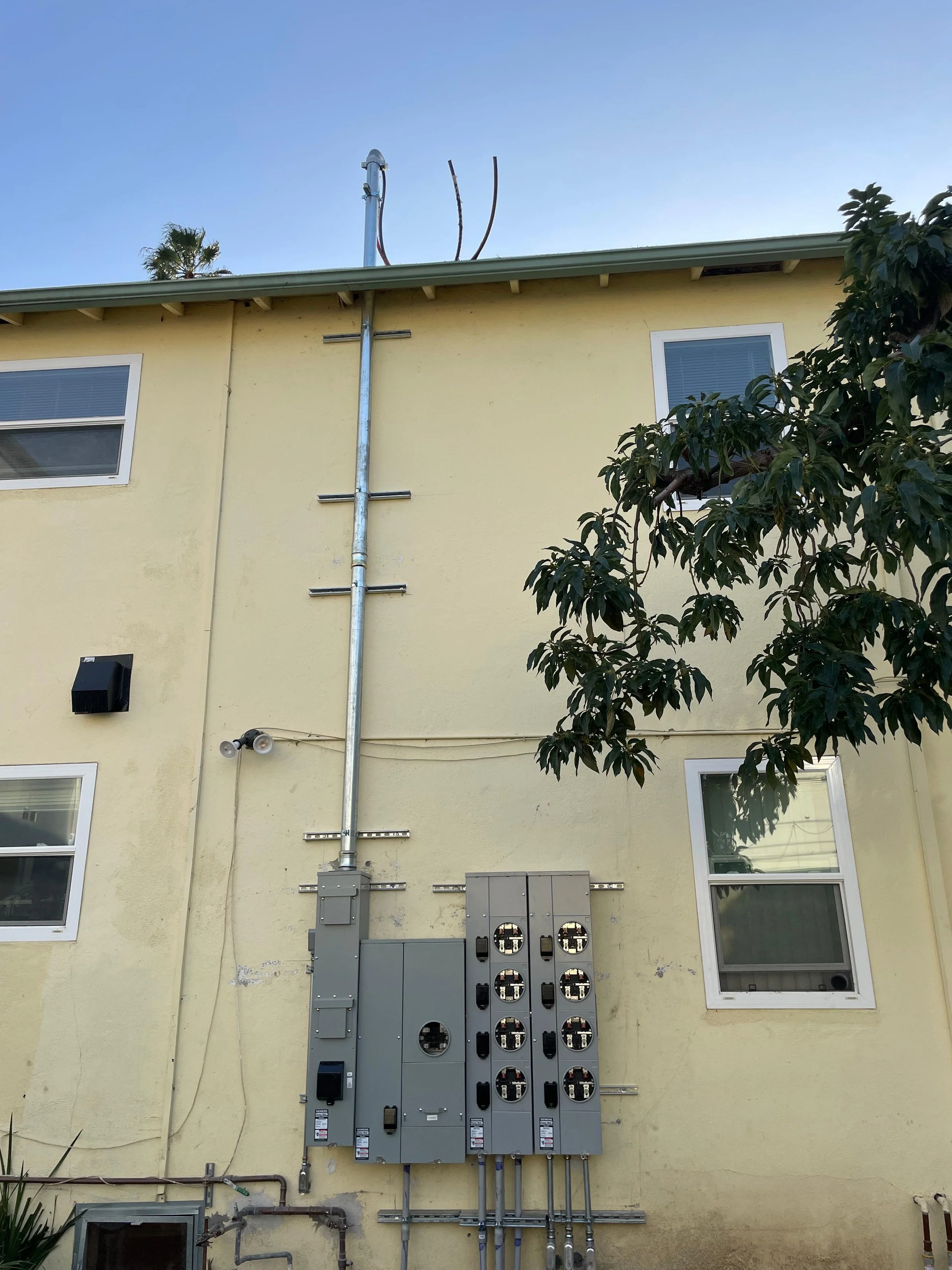 The image shows the exterior of a building with a yellow wall, two white-framed windows, and various electrical and utility boxes. A metal conduit pipe runs vertically along the wall, and there are electrical meters and components mounted beneath it.