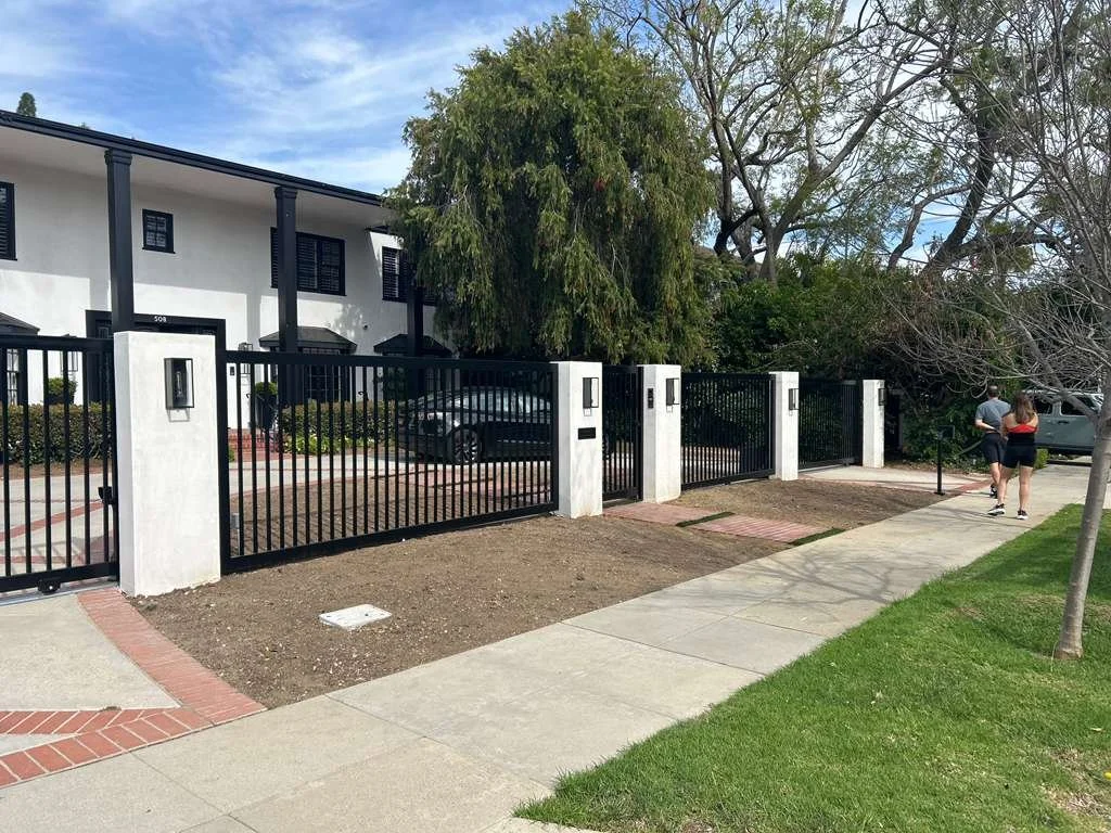 A residential street scene with a gated driveway, a white two-story building in the background, and two people walking on the sidewalk.
