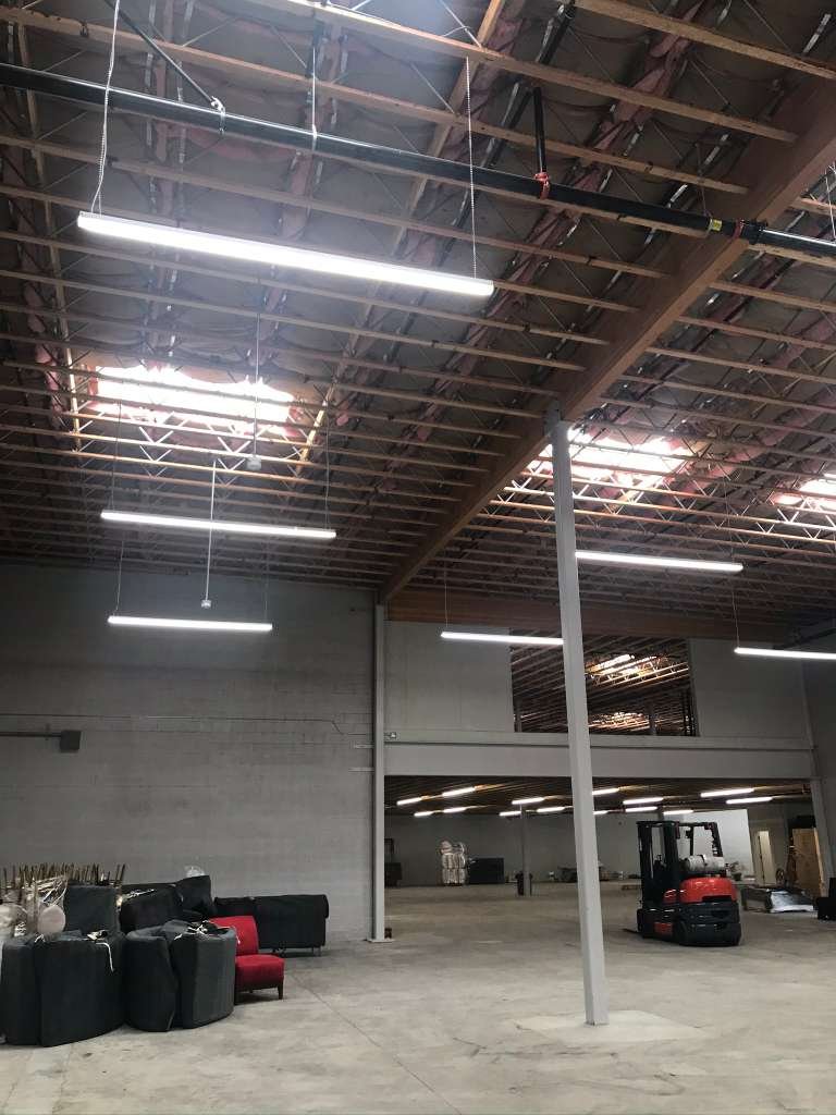 Interior of an unfinished warehouse or industrial space with exposed ceiling beams, hanging fluorescent lights, and warehouse equipment like a forklift and sofas stacked with other furniture.