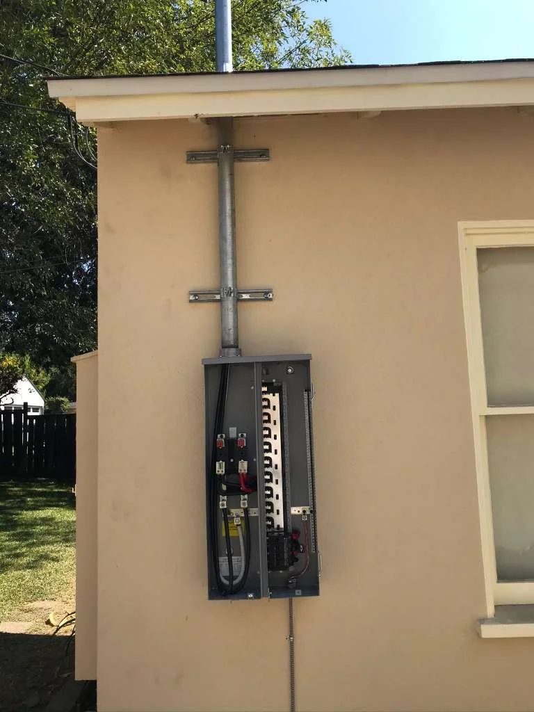 Electric service panel with exposed wiring and conduit outside a beige house, under a roof overhang.