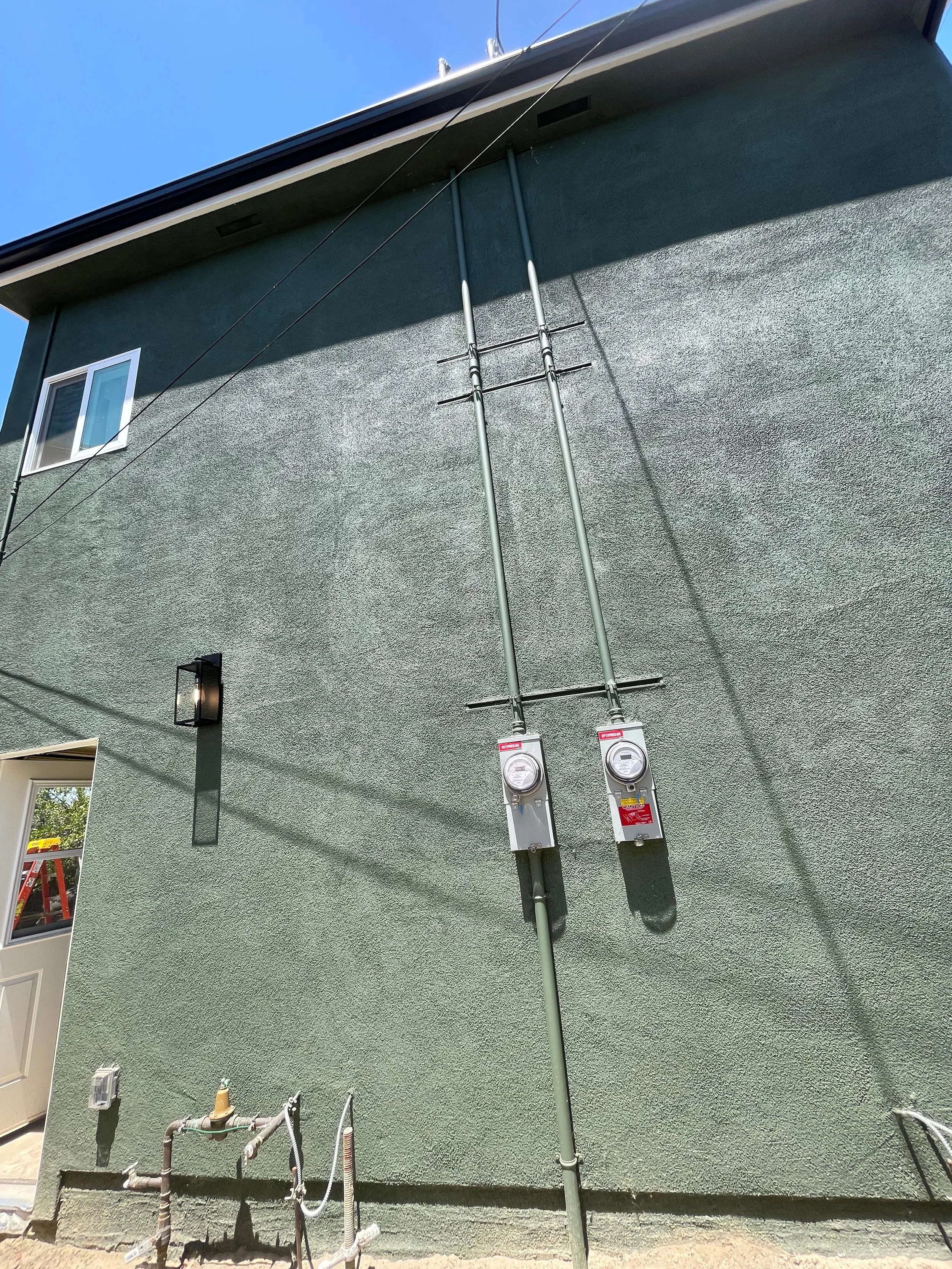 The exterior wall of a house with an aerator and electric meters installed, along with some plumbing fixtures and wires, under a bright blue sky.