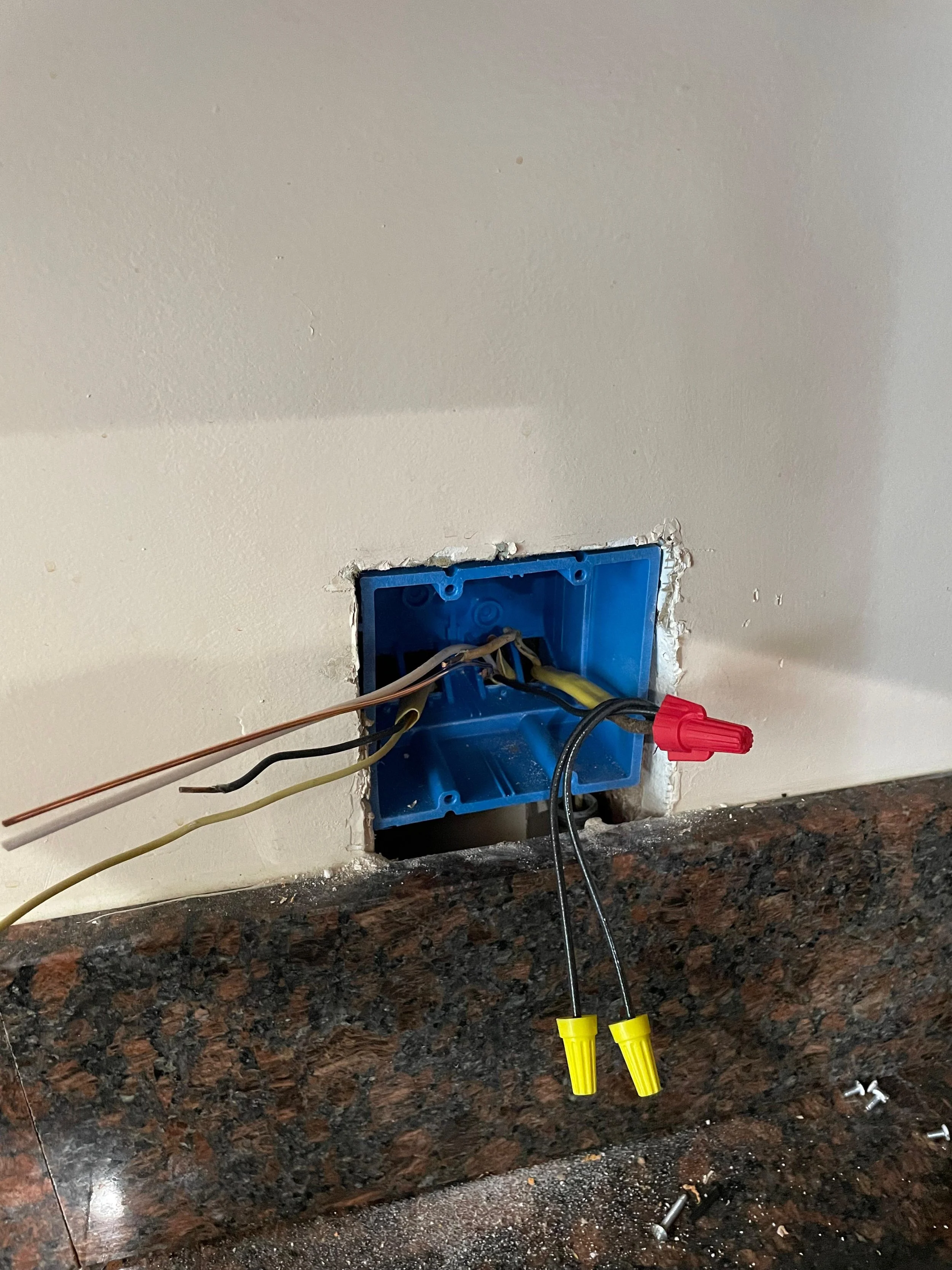 Open electrical outlet box with wires and a yellow test probe hanging in a kitchen wall above a granite countertop.