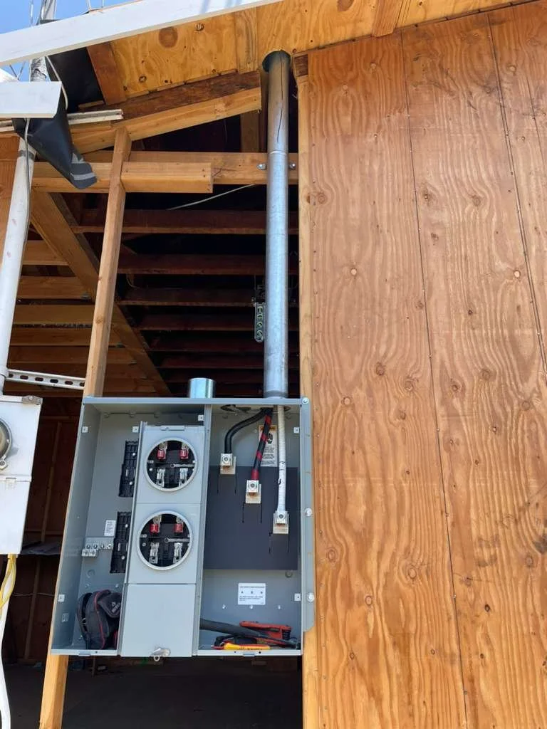 Open electrical panel with wiring, circuit breakers, and tools inside, next to a wooden wall and staircase.