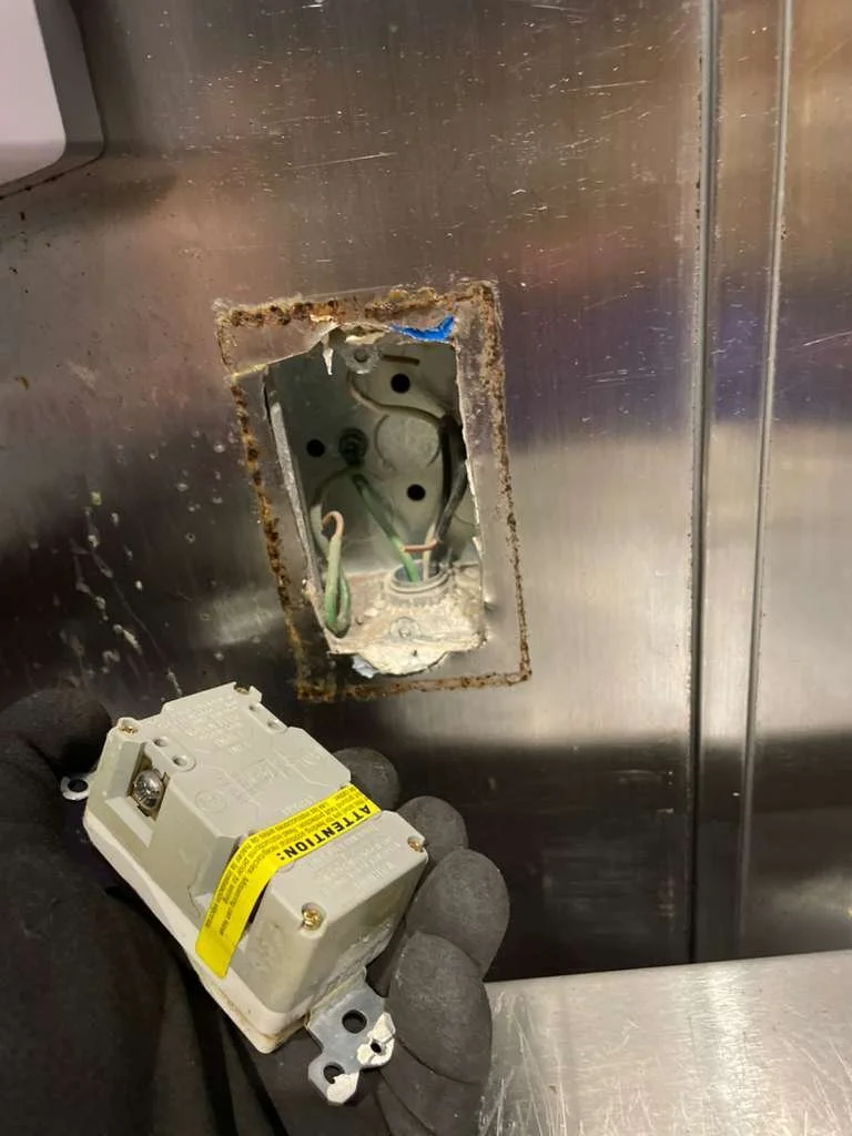 A person wearing black gloves holding a circuit breaker near an electrical outlet with exposed wires and a rusty metal wall.