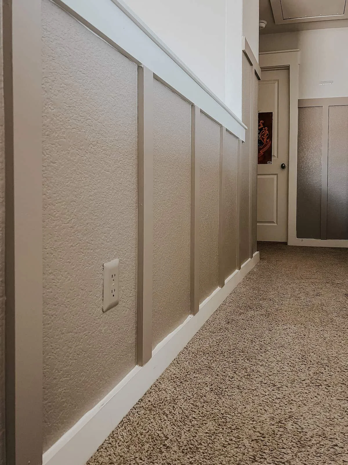 Interior of a room showing a beige textured wall with an electrical outlet, a carpeted floor, and a white door in the background.