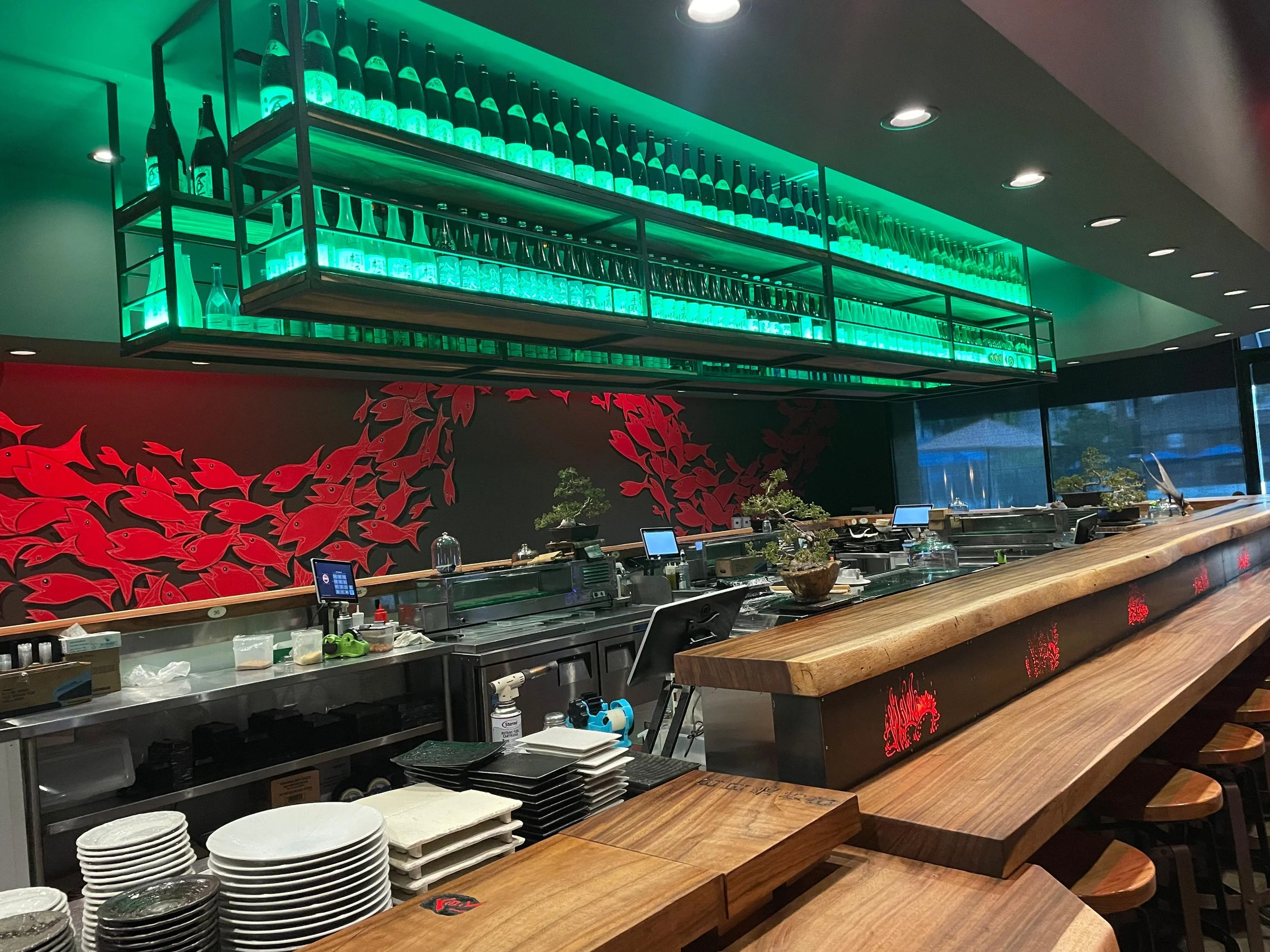 A restaurant or bar counter with plates and utensils, a red fish-themed mural on the wall, and a hanging shelf with bottles and glasses illuminated by green lighting.