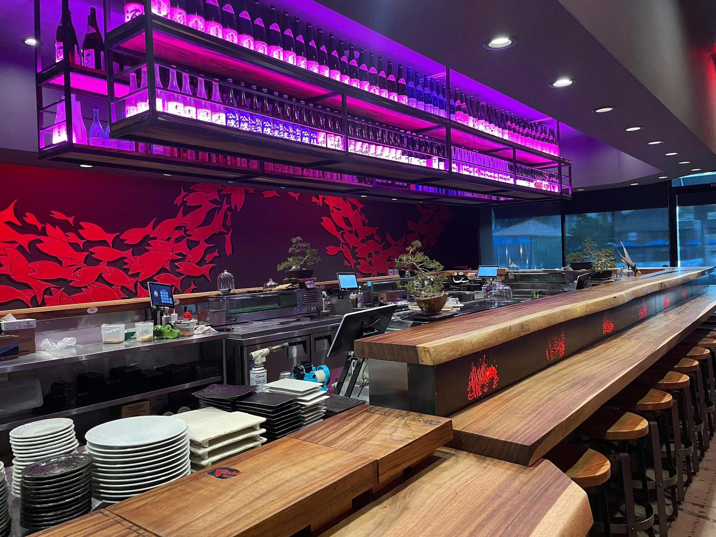 Interior of a restaurant or bar with a long wooden counter, red wall art featuring fish, and a shelf with bottles illuminated by purple lighting, with several plates and utensils on the counter.