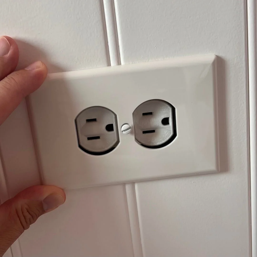 A white electrical outlet with two power sockets on a white wall, with a person's hand holding the cover plate.