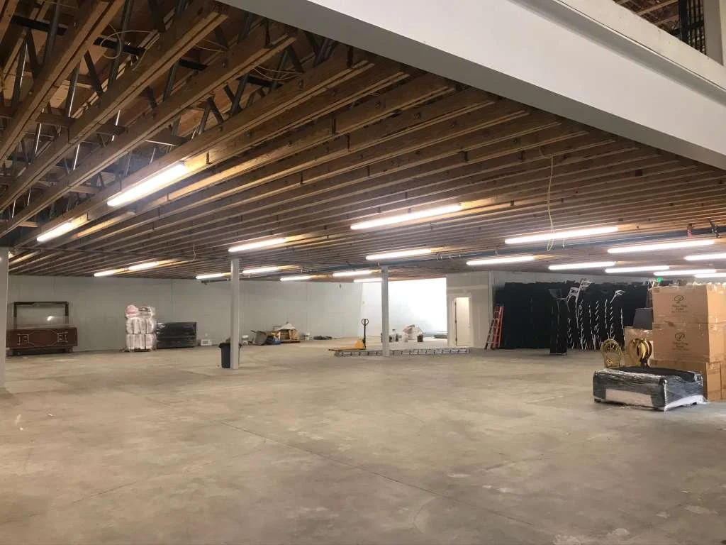 An unfinished indoor space with a wooden ceiling, fluorescent lighting, and construction materials scattered on the concrete floor.