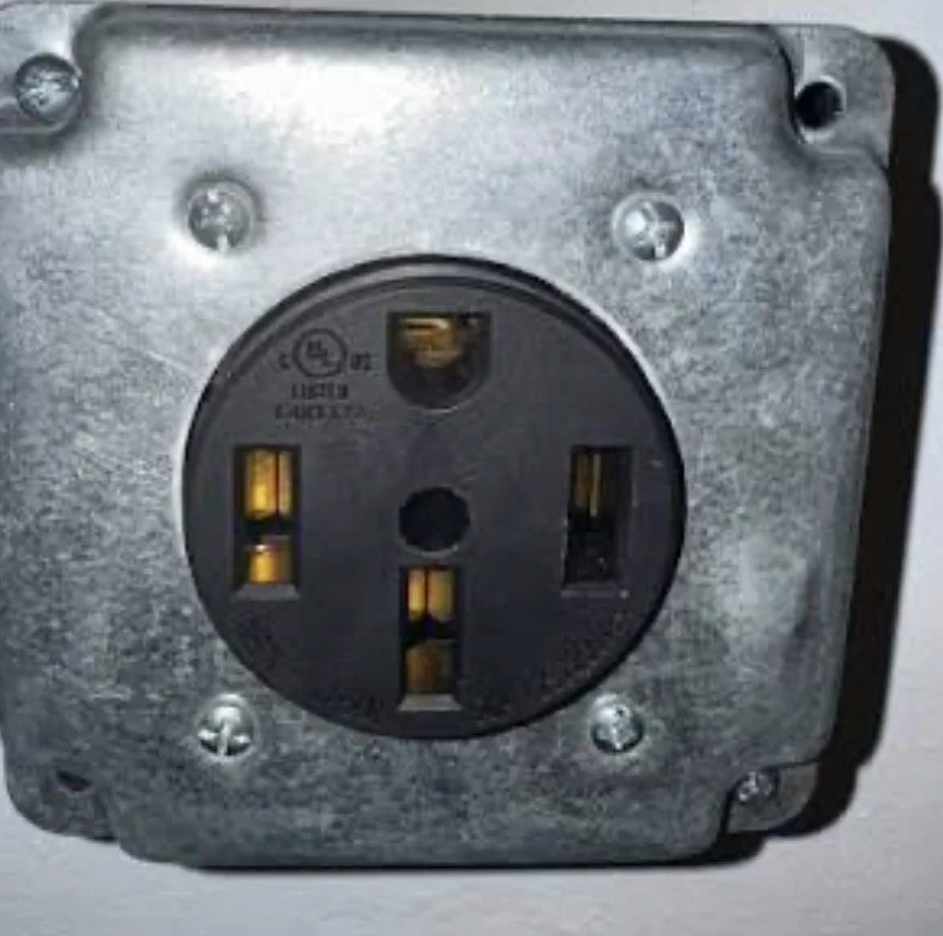 Close-up of a metallic electrical power outlet, with a three-prong socket and screws around the edges.