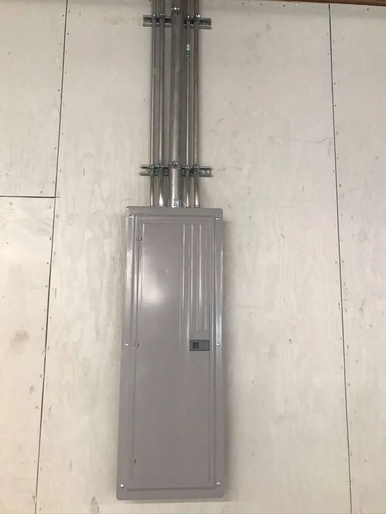 Electrical panel box mounted on a wall with metal conduit pipes running vertically above it.