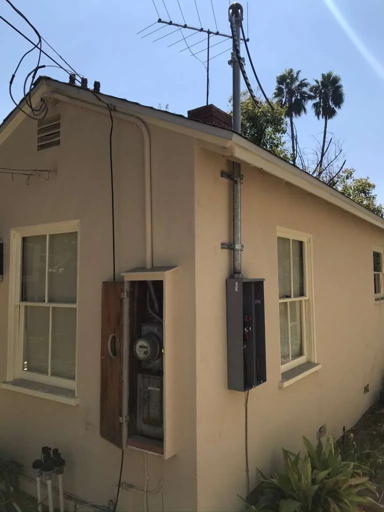 Exterior view of a house with two windows, electrical meter, and electrical panel box on the side wall, with palm trees and power lines in the background.