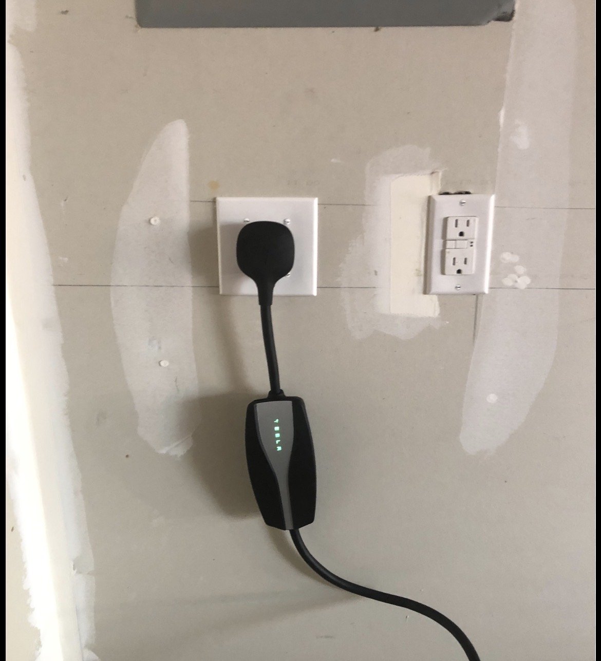Electric vehicle charging cable plugged into a wall outlet, with a charging station display showing progress, on a wall with unfinished drywall.