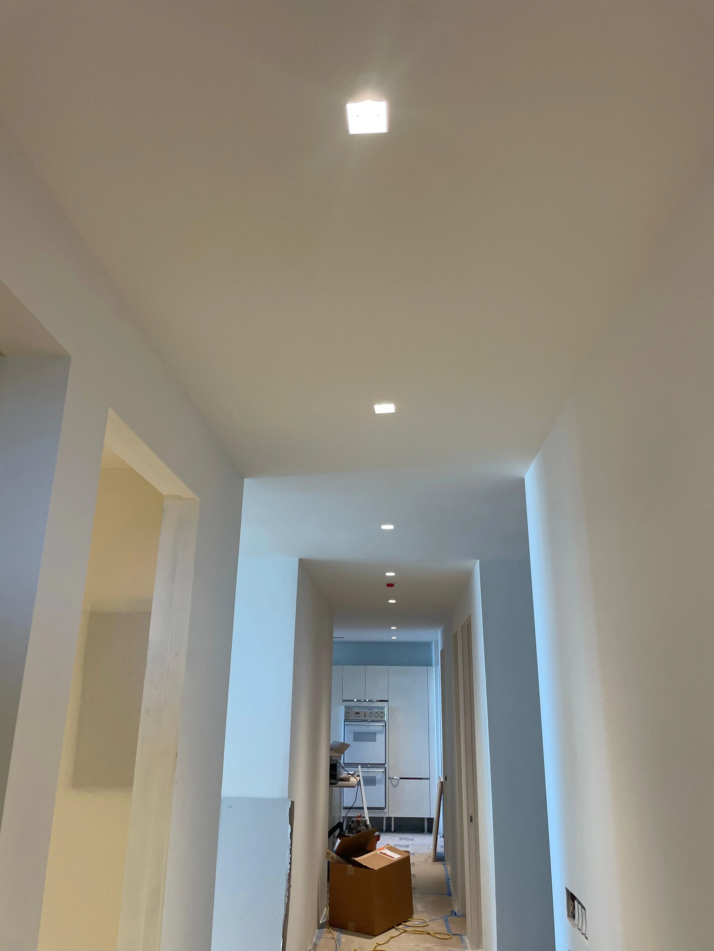 A hallway under construction with white walls and ceiling, opening into a kitchen with white cabinetry and appliances, and construction tools and a cardboard box on the floor.