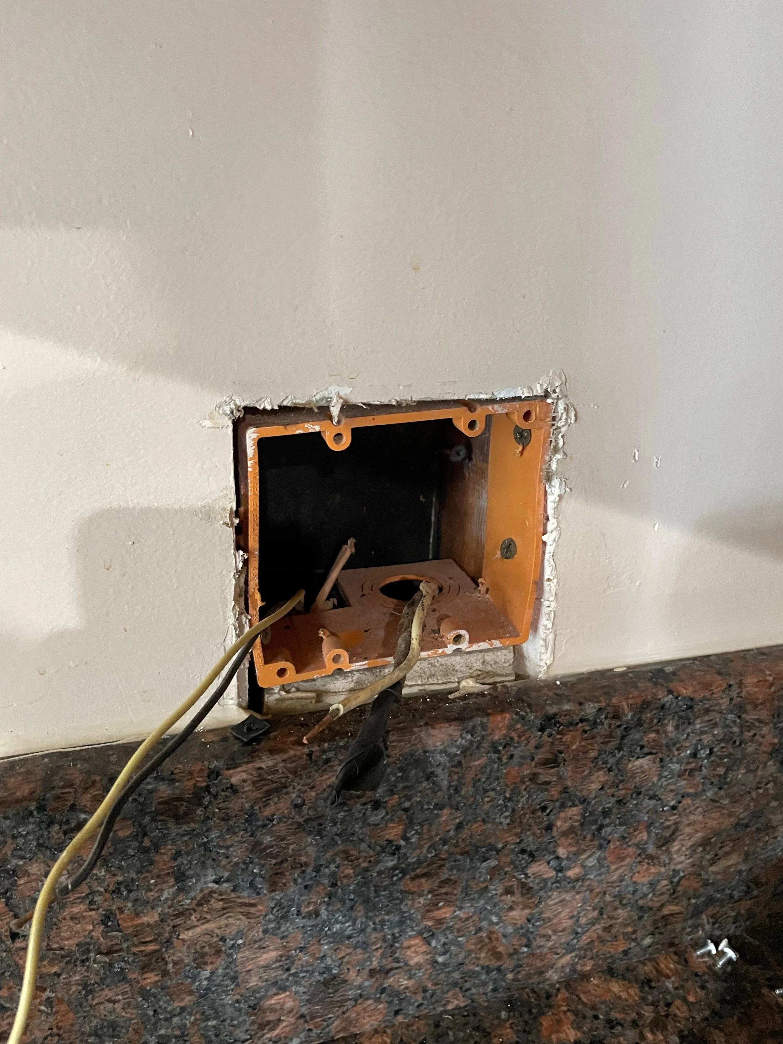 Open electrical outlet box with exposed wires on a white wall above a dark granite countertop.