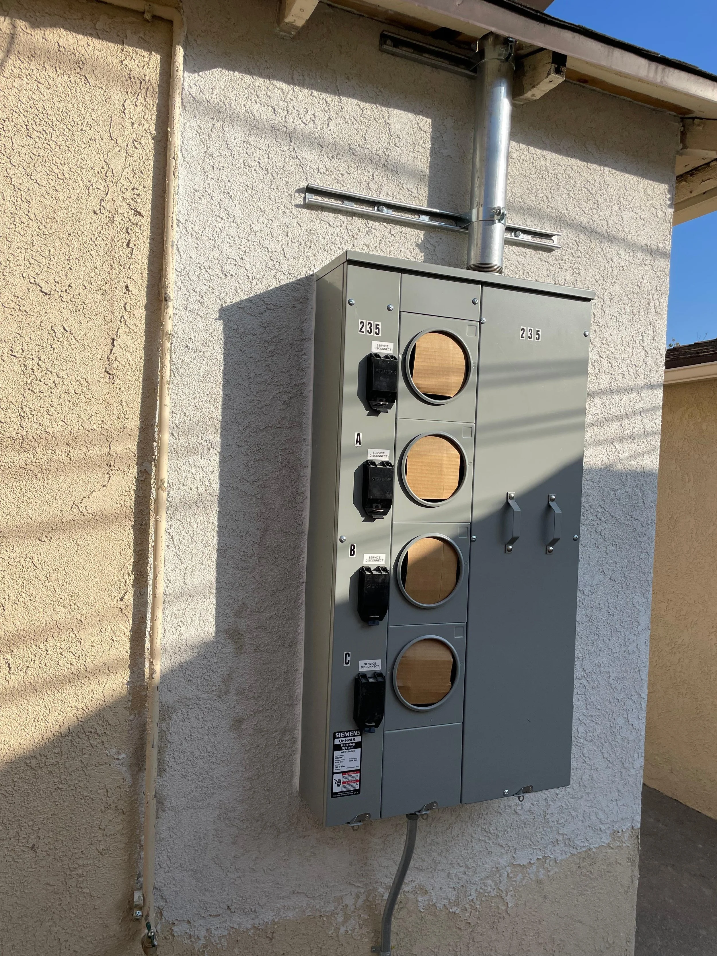An outdoor electrical meter box installed on a stucco wall, with three circular cut-outs covered by wooden boards and labeled sections A, B, and C.