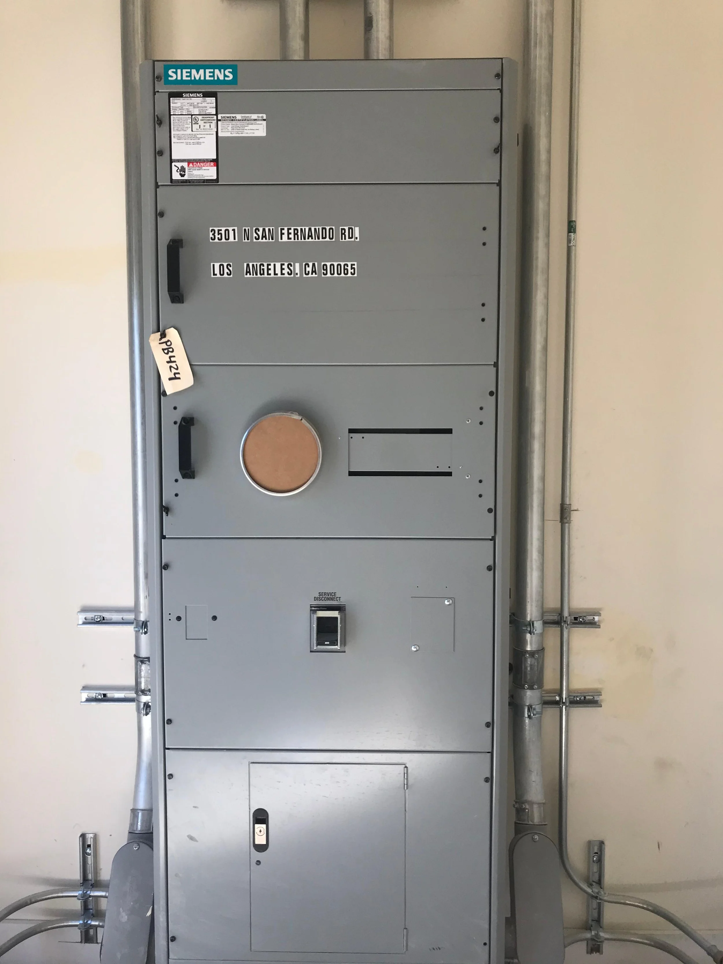 Gray electrical panel with Siemens branding, several labels and switches, installed on a wall with visible metal conduits.