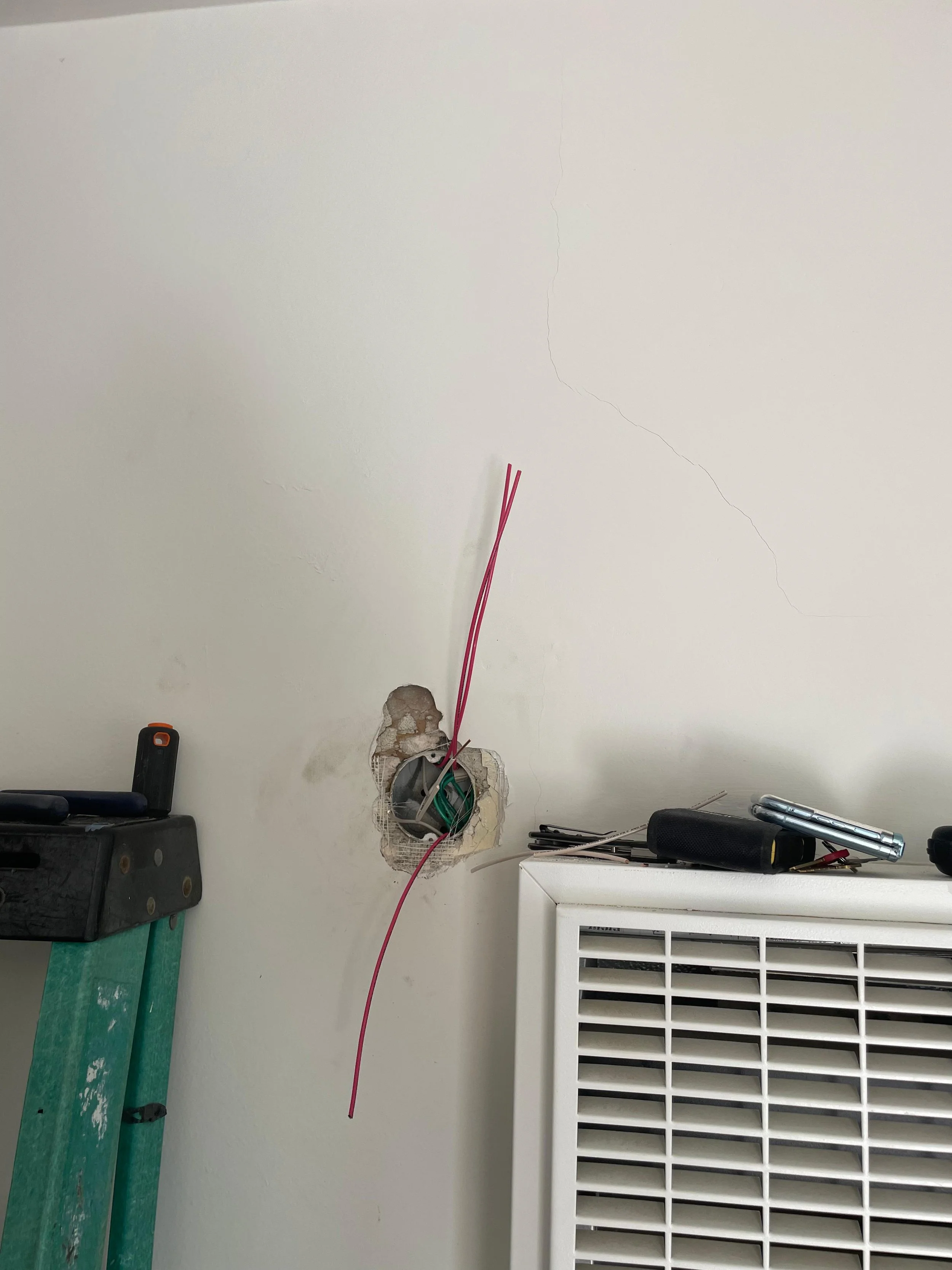 Construction or repair work involving electrical wiring in a wall. The wall has a hole with exposed wiring, and there are tools and a vent or heater below.
