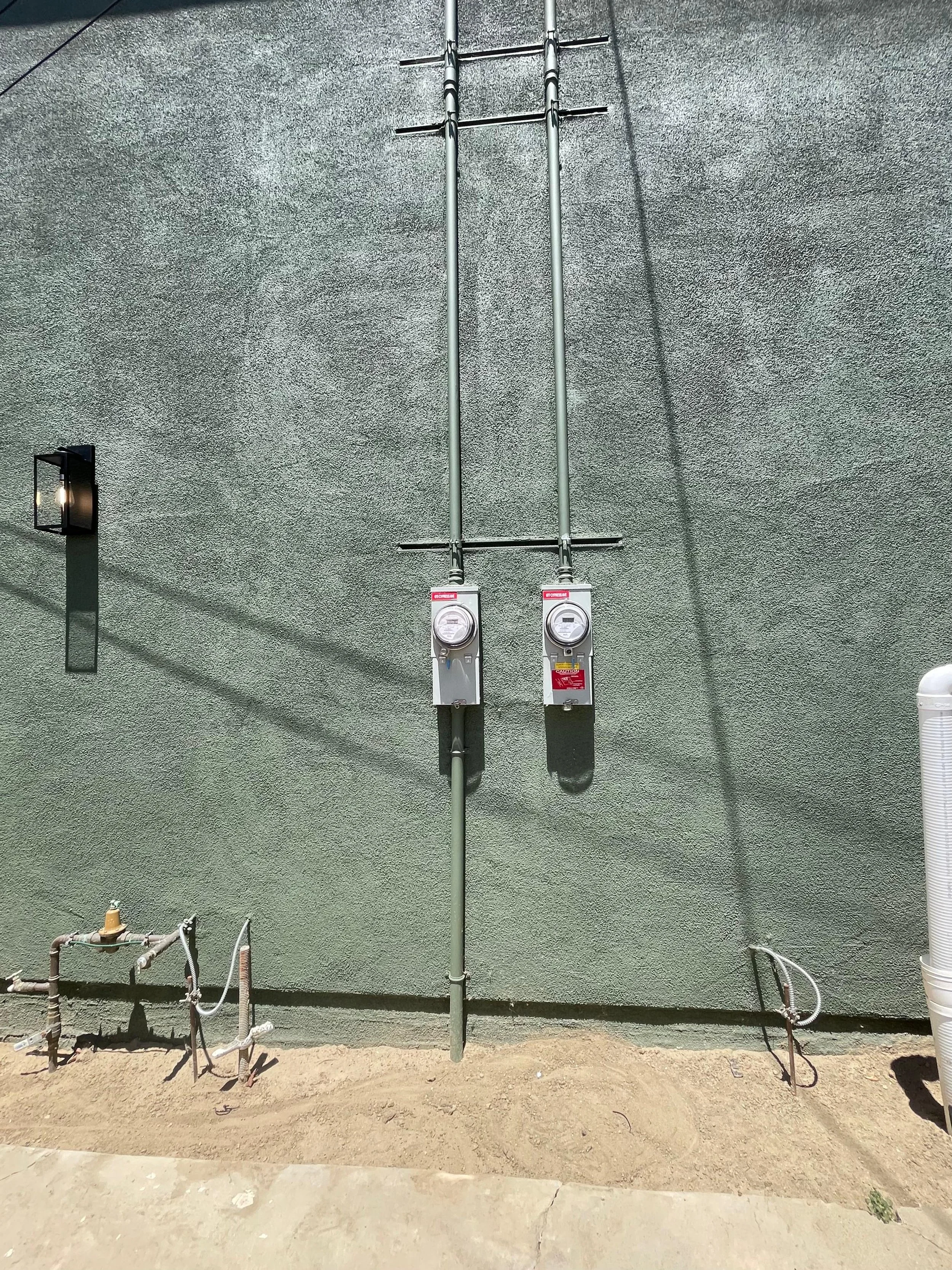 Electrical meters and conduit pipes mounted on a textured gray wall outside a building, with utility boxes and piping at the ground level.