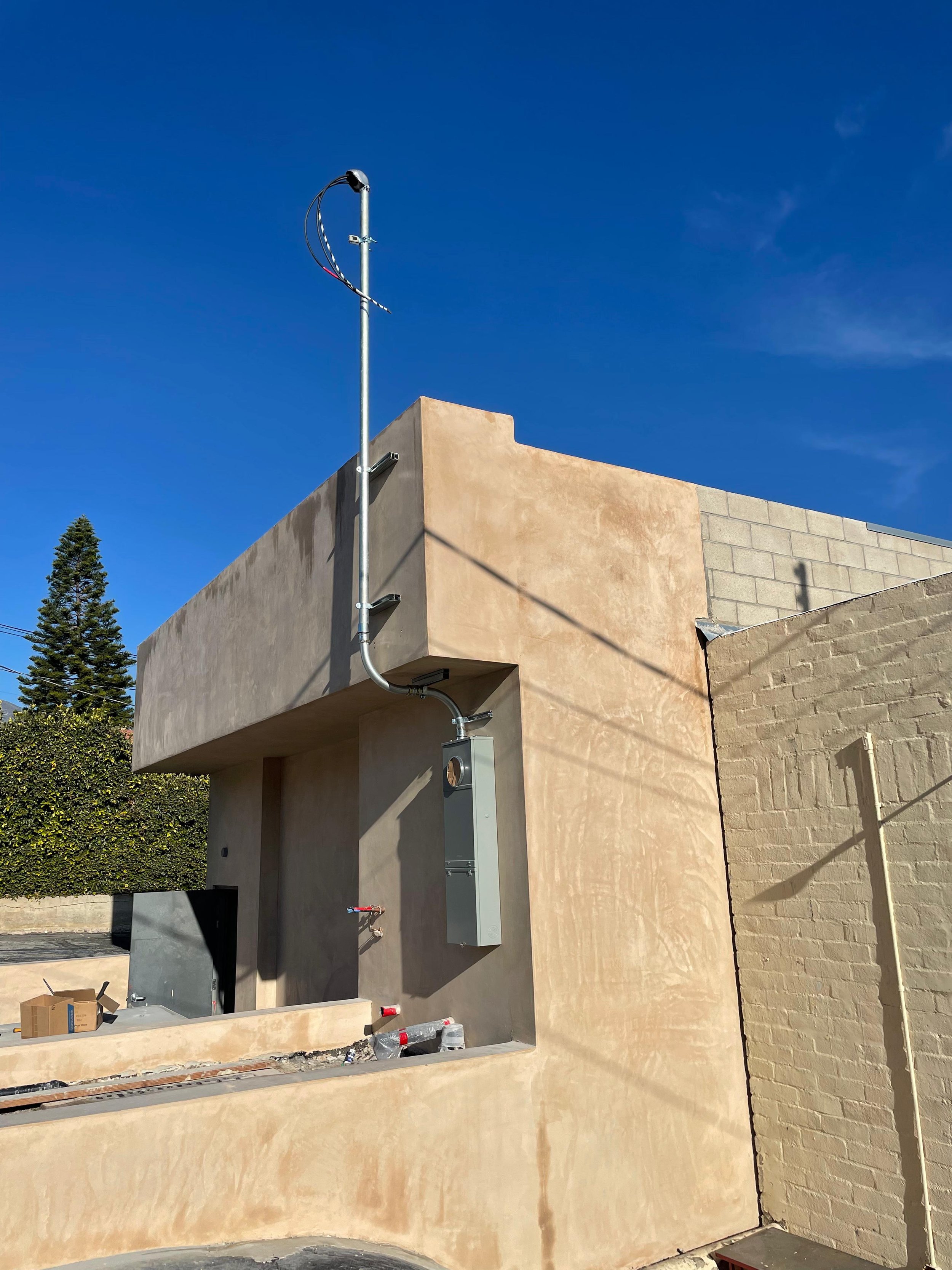 Exterior of a building under construction or renovation with unfinished walls and exposed piping, against a clear blue sky. There are construction supplies and equipment visible, including a gray electrical box, a box of tools, and a cardboard box.