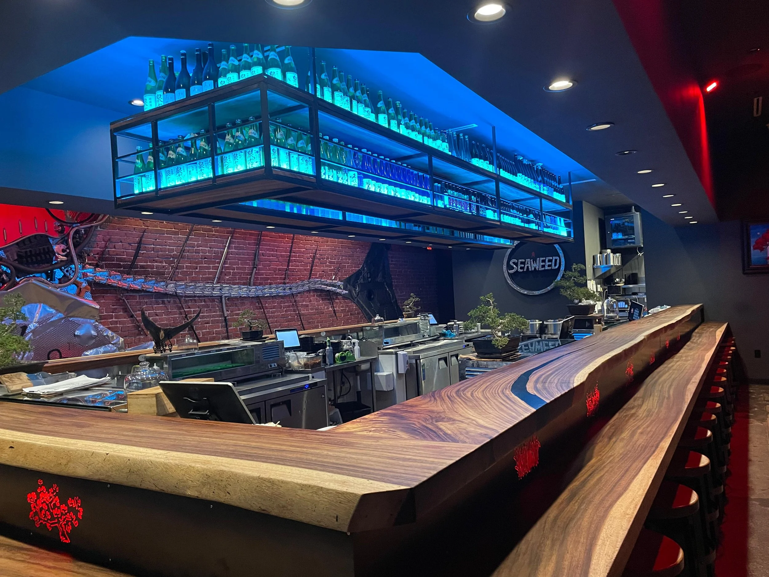Bar area with bottles of sake displayed on illuminated shelves above the counter, and a red Chinese character logo on the side of the counter.