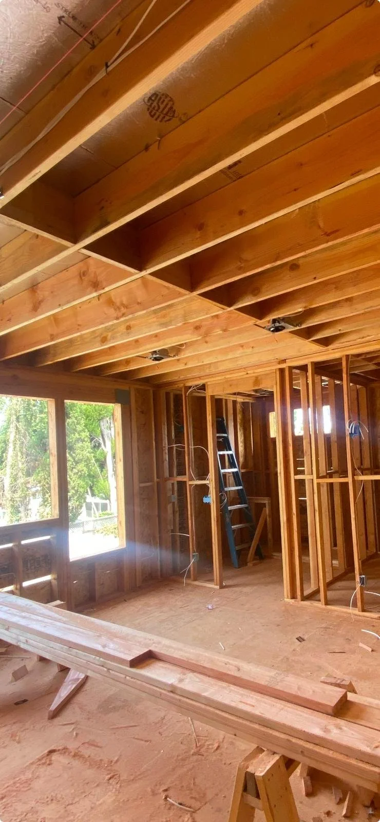 Interior view of a house under construction showing exposed wooden framing, ceiling beams, electrical wiring, and a ladder, with a large window letting in natural light.