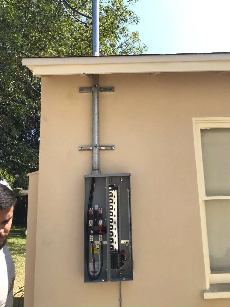 Electrical service panel with conduit and wiring mounted on the exterior wall of a building, partially visible person on the left, window on the right.