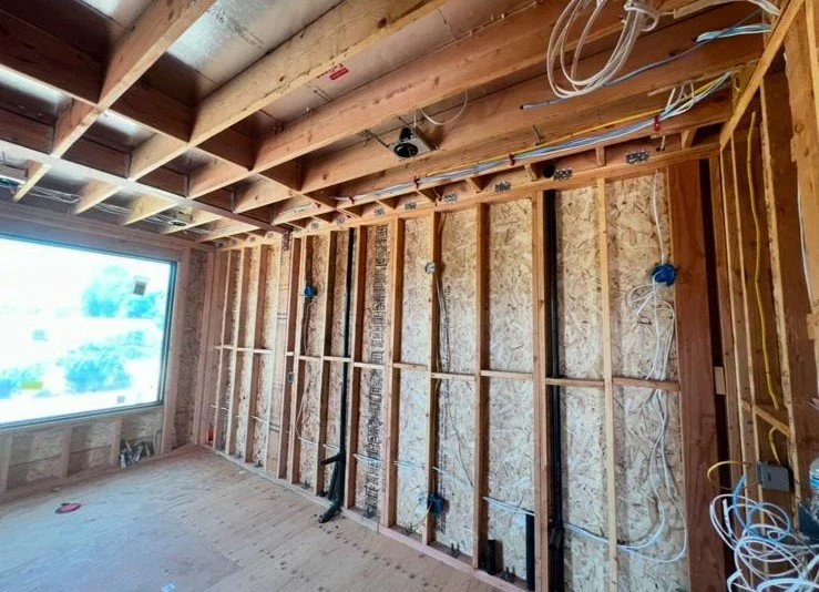 Construction site of an unfinished room with exposed wooden studs and ceiling, electrical wiring installed, and a large window letting in natural light.