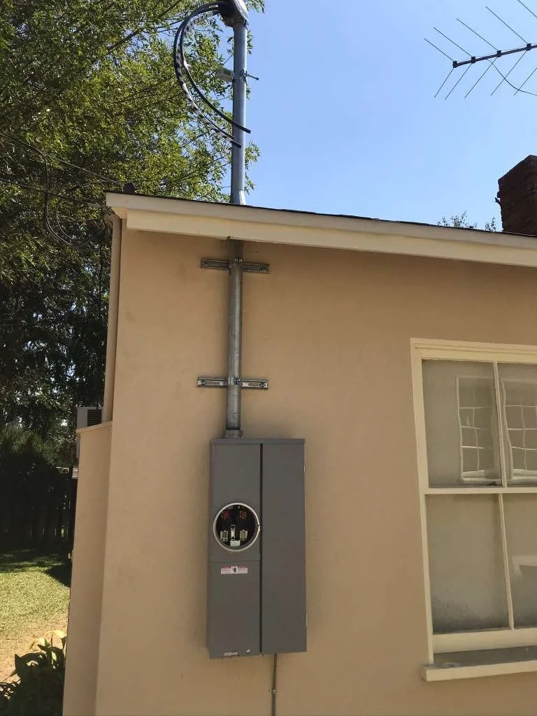 Exterior wall of a house with an electric meter box and power meter on it. Several wires are connected to the meter, and a tall antenna is mounted on the roof.