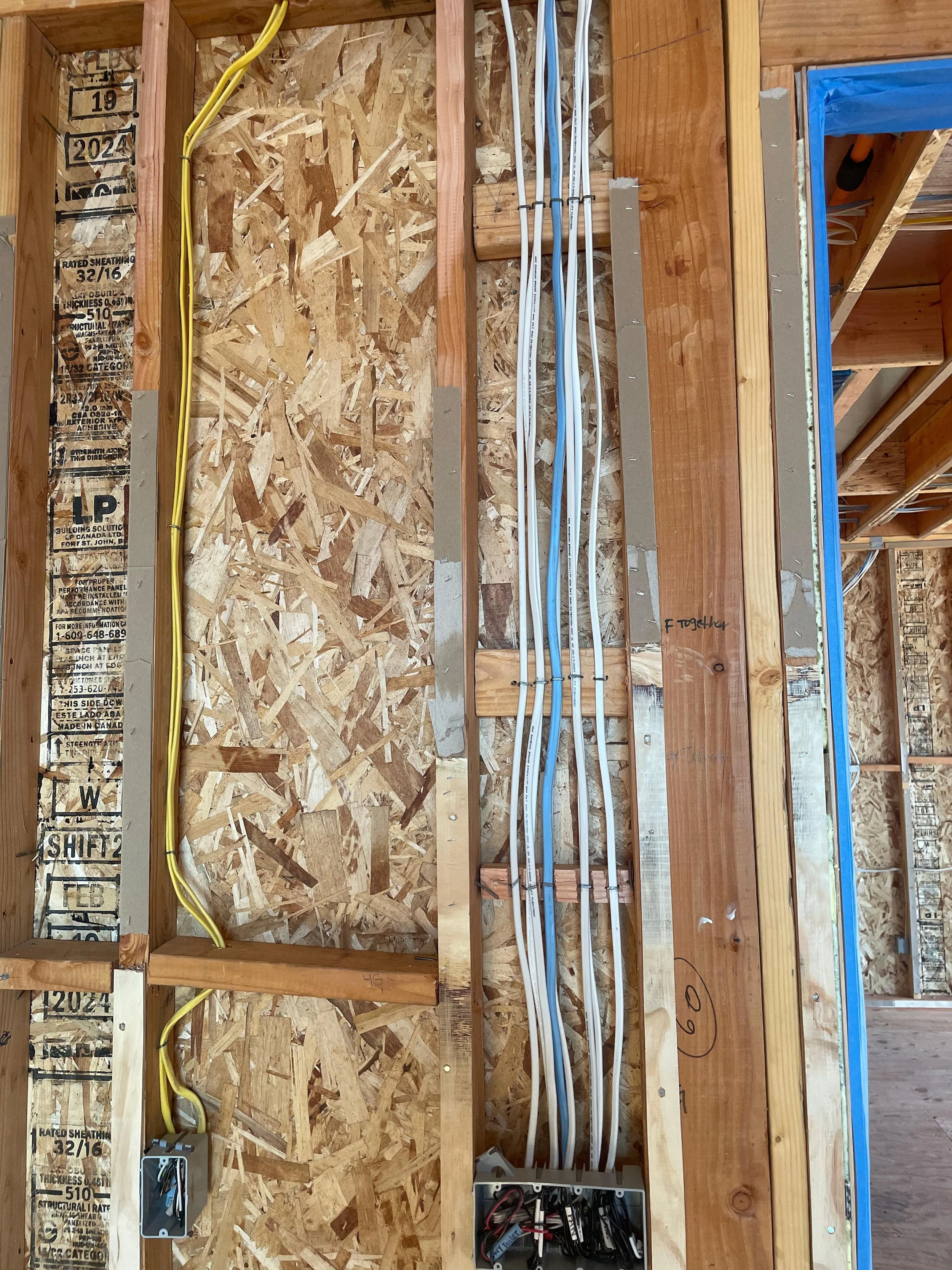 Electrical wiring and outlets on a construction site wall, with yellow and blue electrical cables, switches, and an electrical junction box installed among unfinished wooden framing and OSB sheathing.