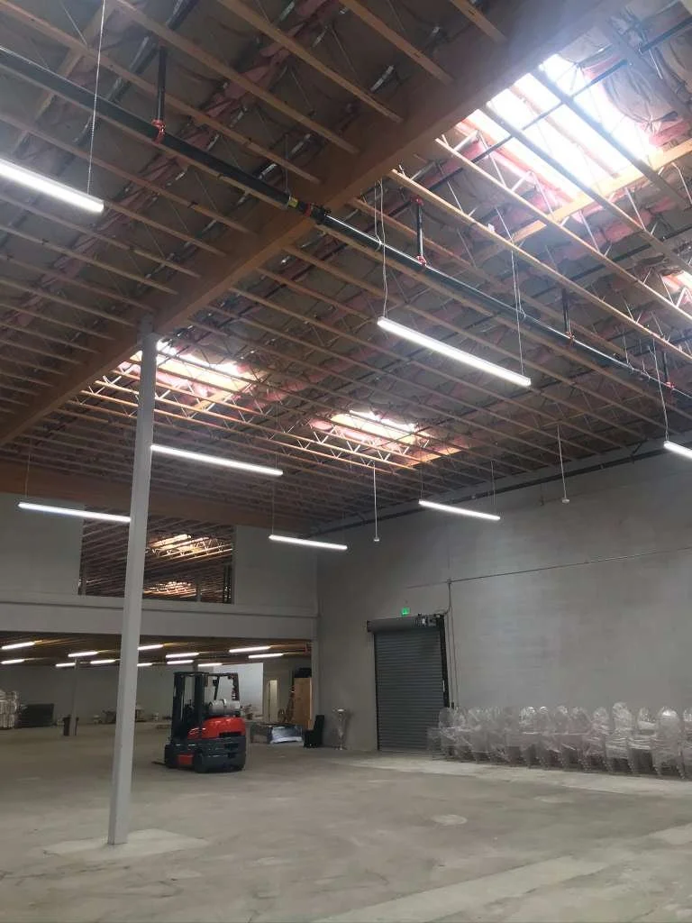 Interior of a warehouse or industrial space with a high ceiling, exposed beams, and skylights, with a forklift and stacked chairs or similar objects along the wall.