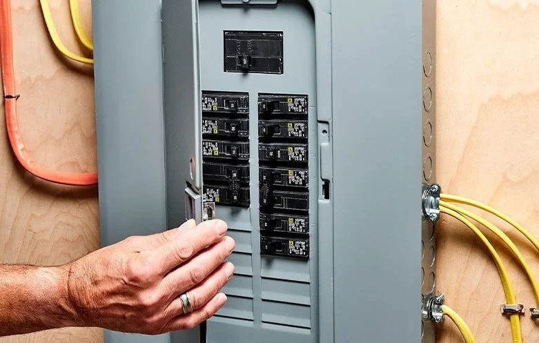 A person is flipping a circuit breaker switch in an electrical panel with several circuit breakers, mounted on a beige wall with yellow and orange wires connected to it.