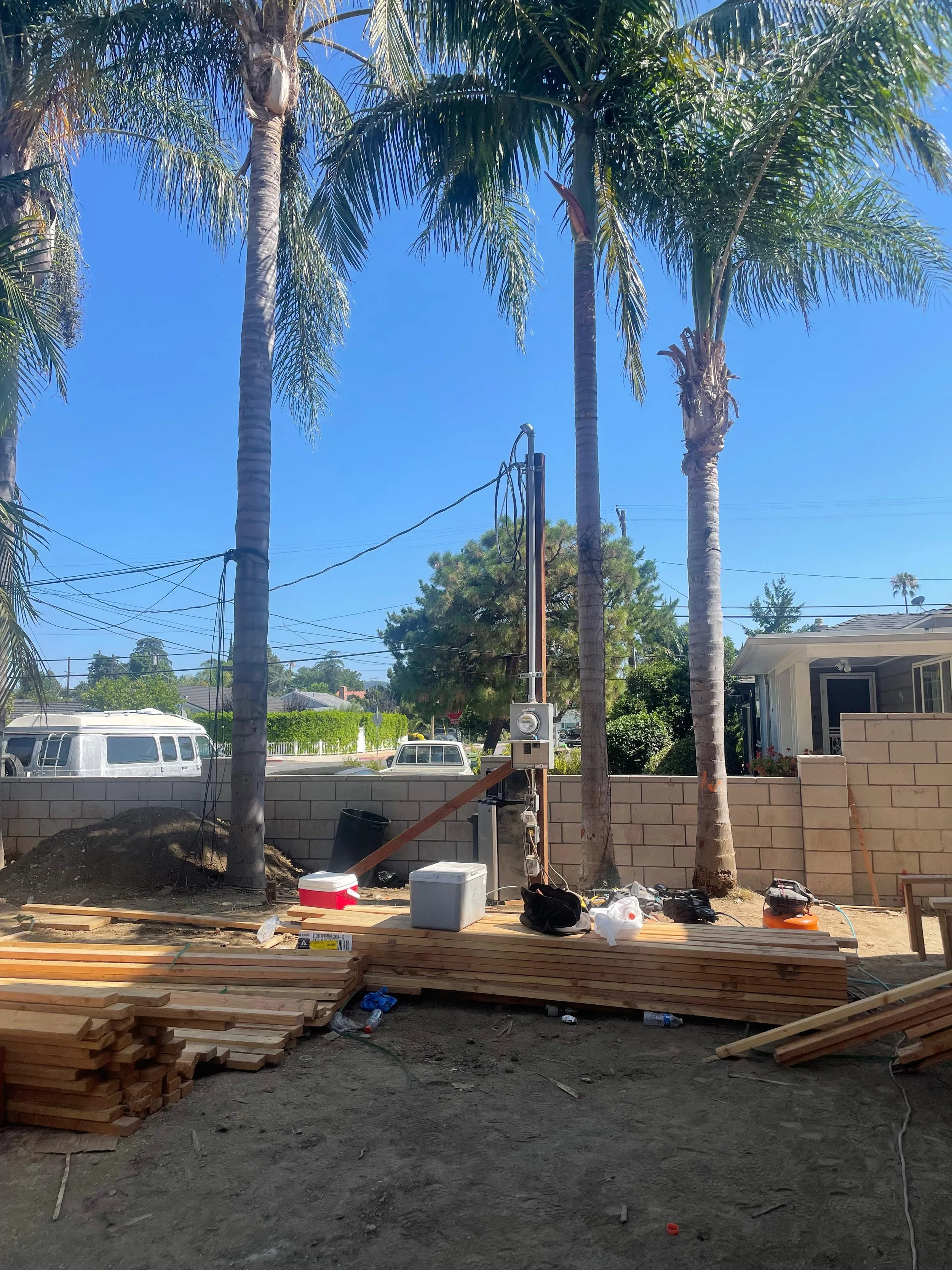 Construction site with several tall palm trees, wooden planks, tools, and construction materials scattered on the ground. A brick wall and parked cars are visible in the background under a clear blue sky.