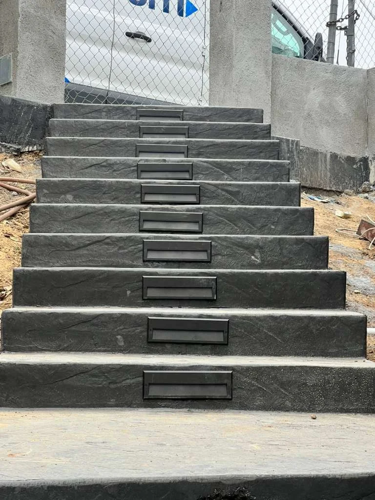 Concrete staircase with eight steps, each having a rectangular black metal insert in the middle of the front face, outside a construction area with soil and debris at the sides and a chain-link fence in the background.