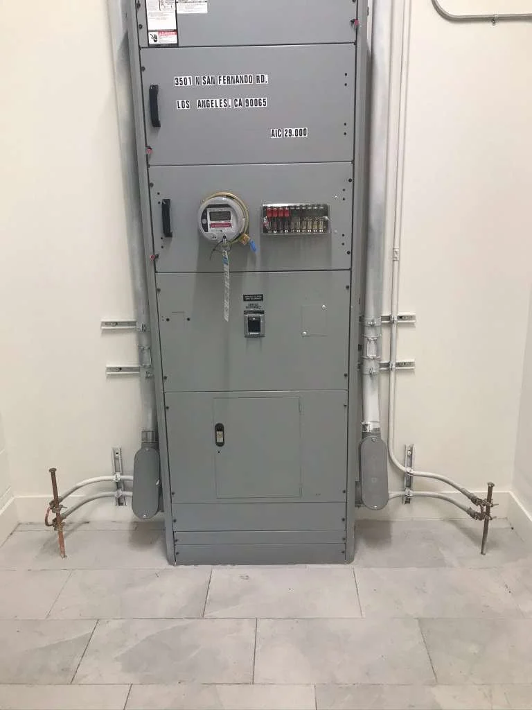 Electrical panel with meters and wiring in a room with tiled floor and white walls.