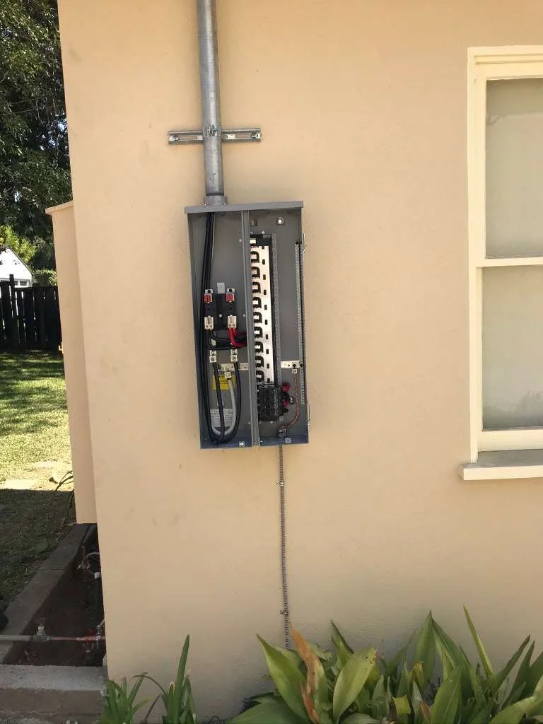 An open electrical box mounted on a beige exterior wall with connected black and red wires, a metal conduit extending upward, and a flexible metal conduit descending downward near some green plants.