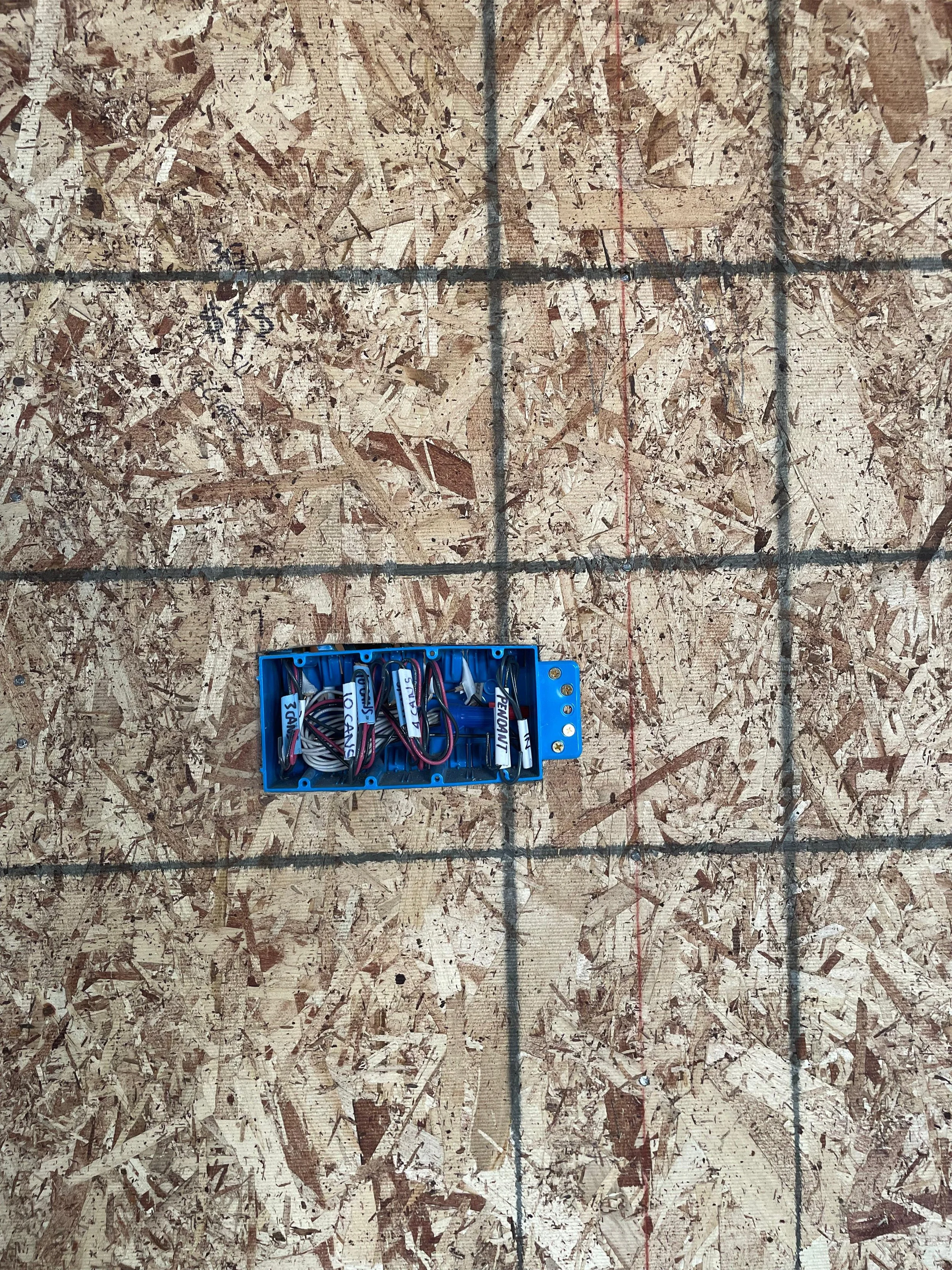 Close-up view of electrical wiring inside a small blue plastic box mounted on a plywood surface with grid markings.