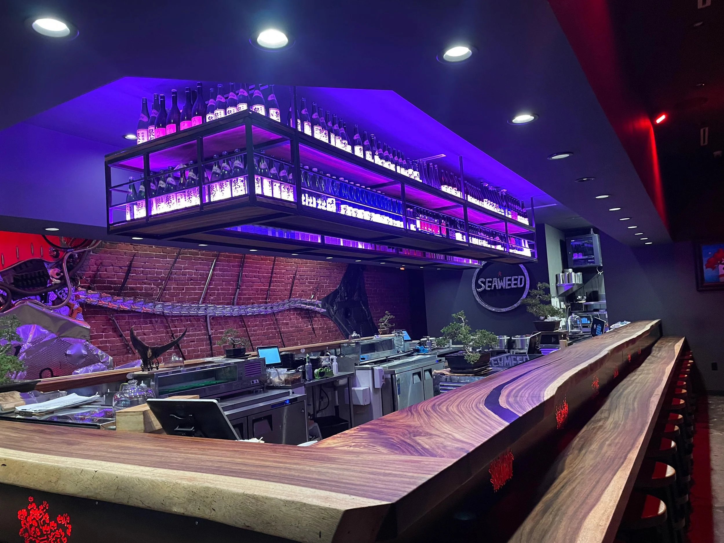 A bar with a curved wooden counter and a high shelf with numerous bottles illuminated with purple lighting. The ceiling has recessed lighting, and there's a brick wall in the background. 