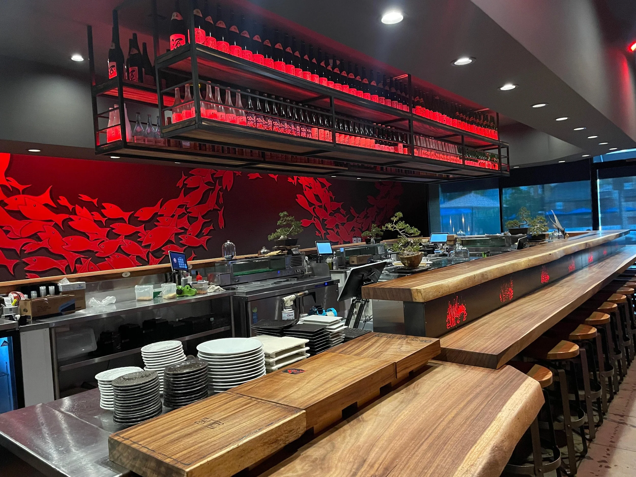 Interior of a restaurant with a wooden bar counter, stacked dishes, and a decorative wall featuring red fish artwork. Shelves above hold bottles and glasses, with red lighting accents.
