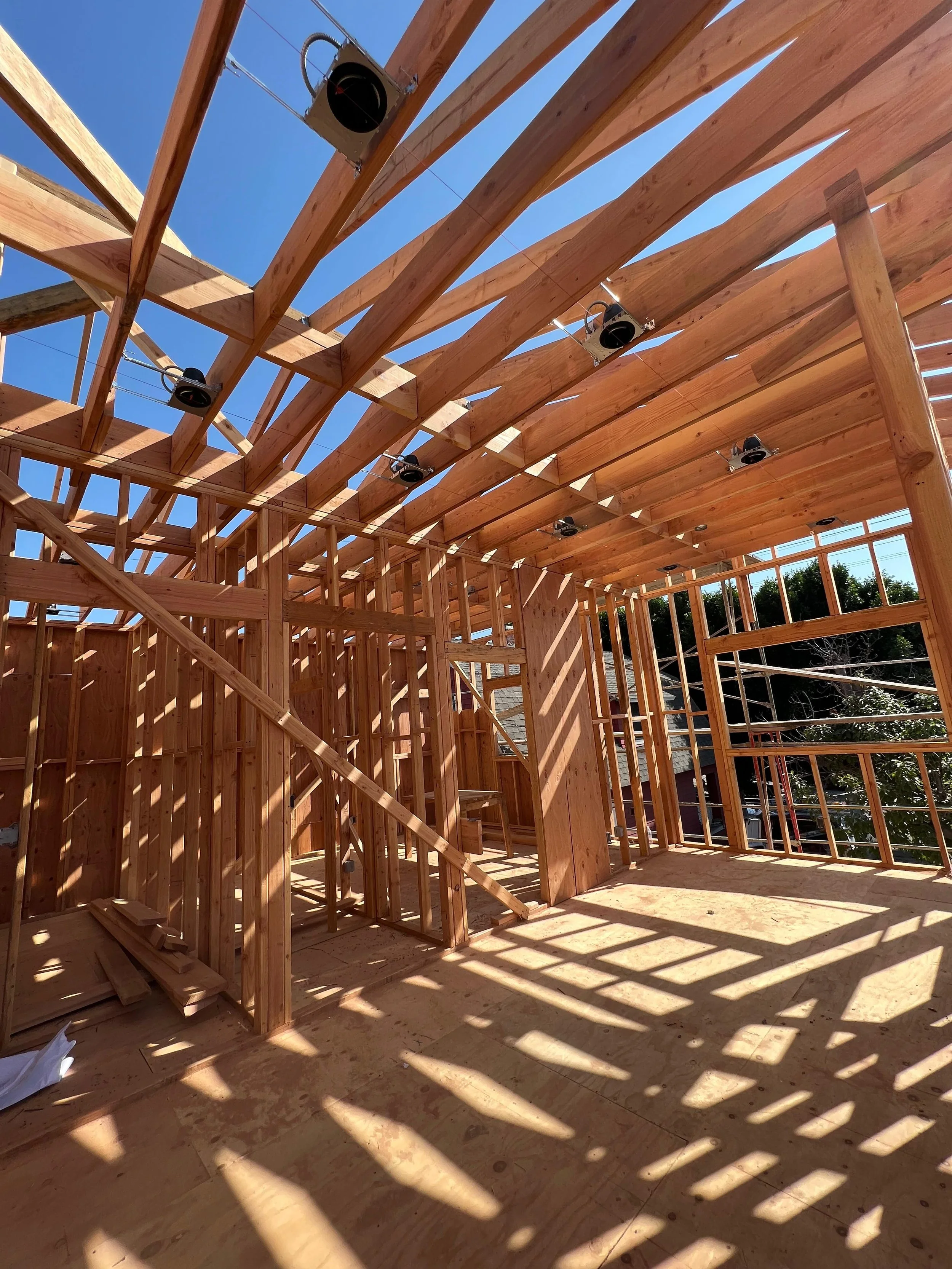 The image shows the wooden framework of a building under construction, with sunlight casting shadows through the open walls and ceiling.