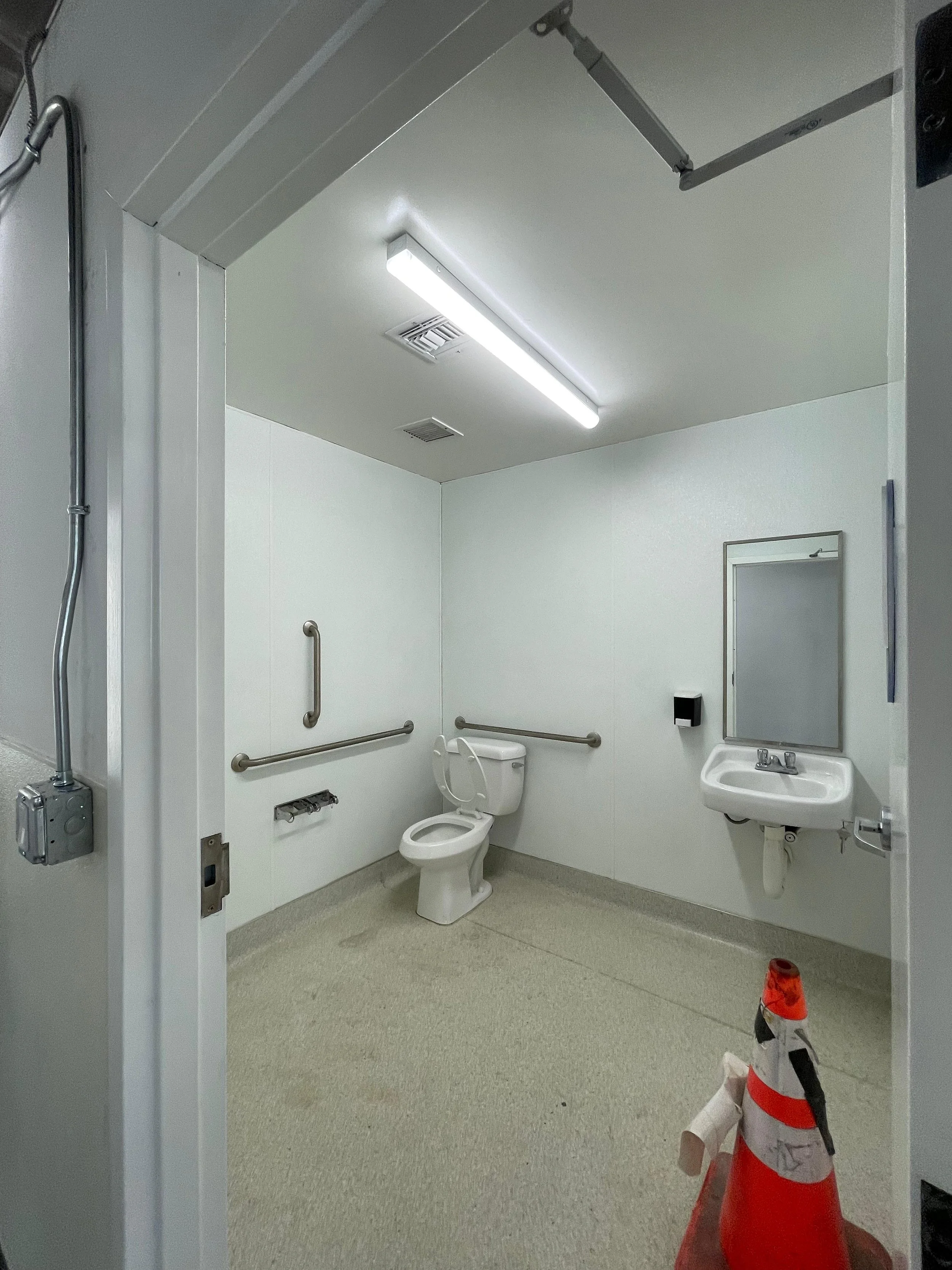 A clean, sterile bathroom with a toilet, small sink, mirror, handrails, and a capped pipe on the floor. There is an orange and white caution cone near the bottom right corner, and a partially open door on the right.