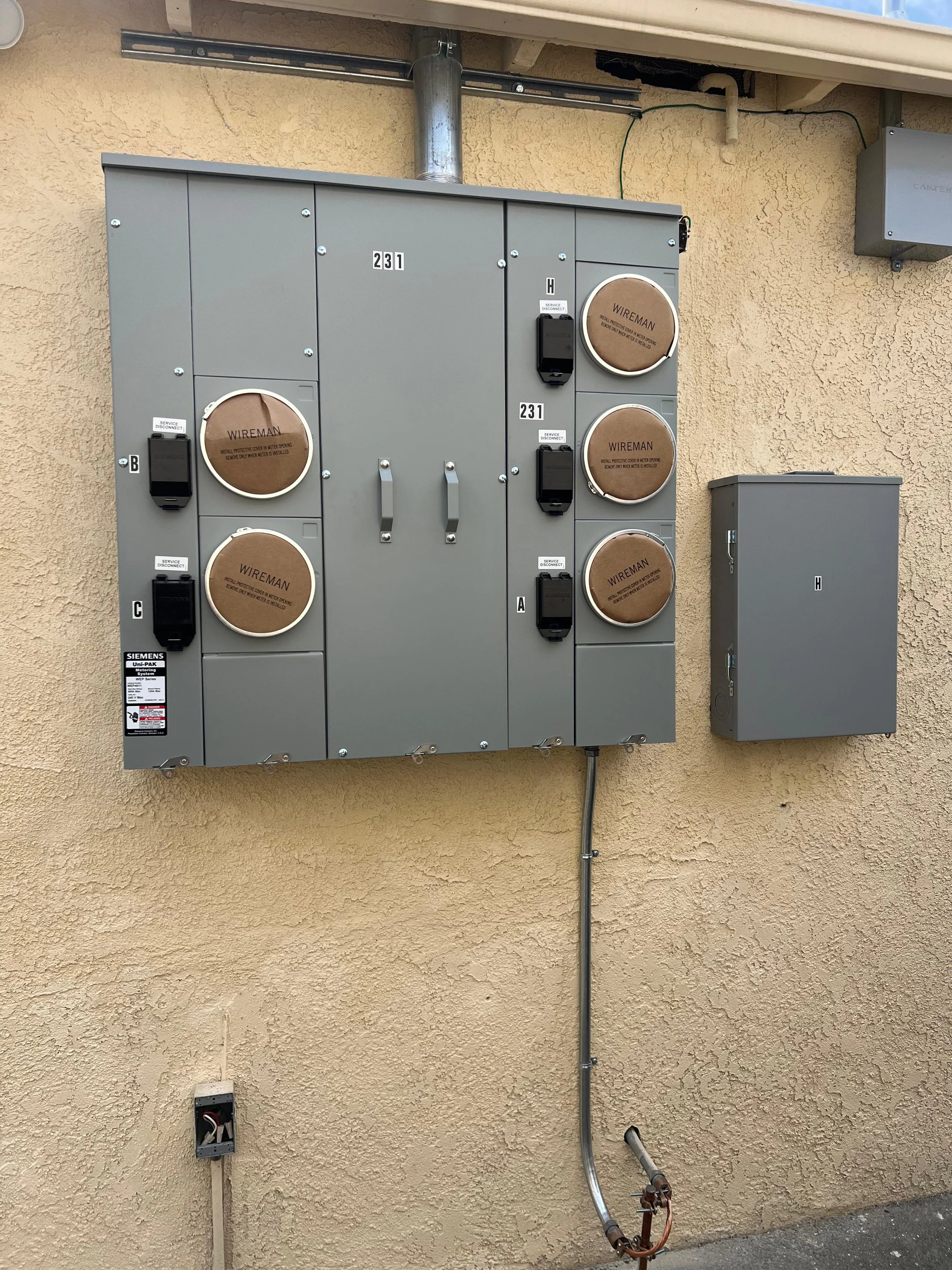 Electrical installation with grey metal electrical boxes mounted on a beige stucco wall. The setup includes multiple circuit boxes labeled with letters and numbers, some covered with brown round covers marked 'WIREMAN.' There are switches labeled 'SERVICE DISCONNECT' and an electrical conduit running vertically from the bottom of the setup to the ground.