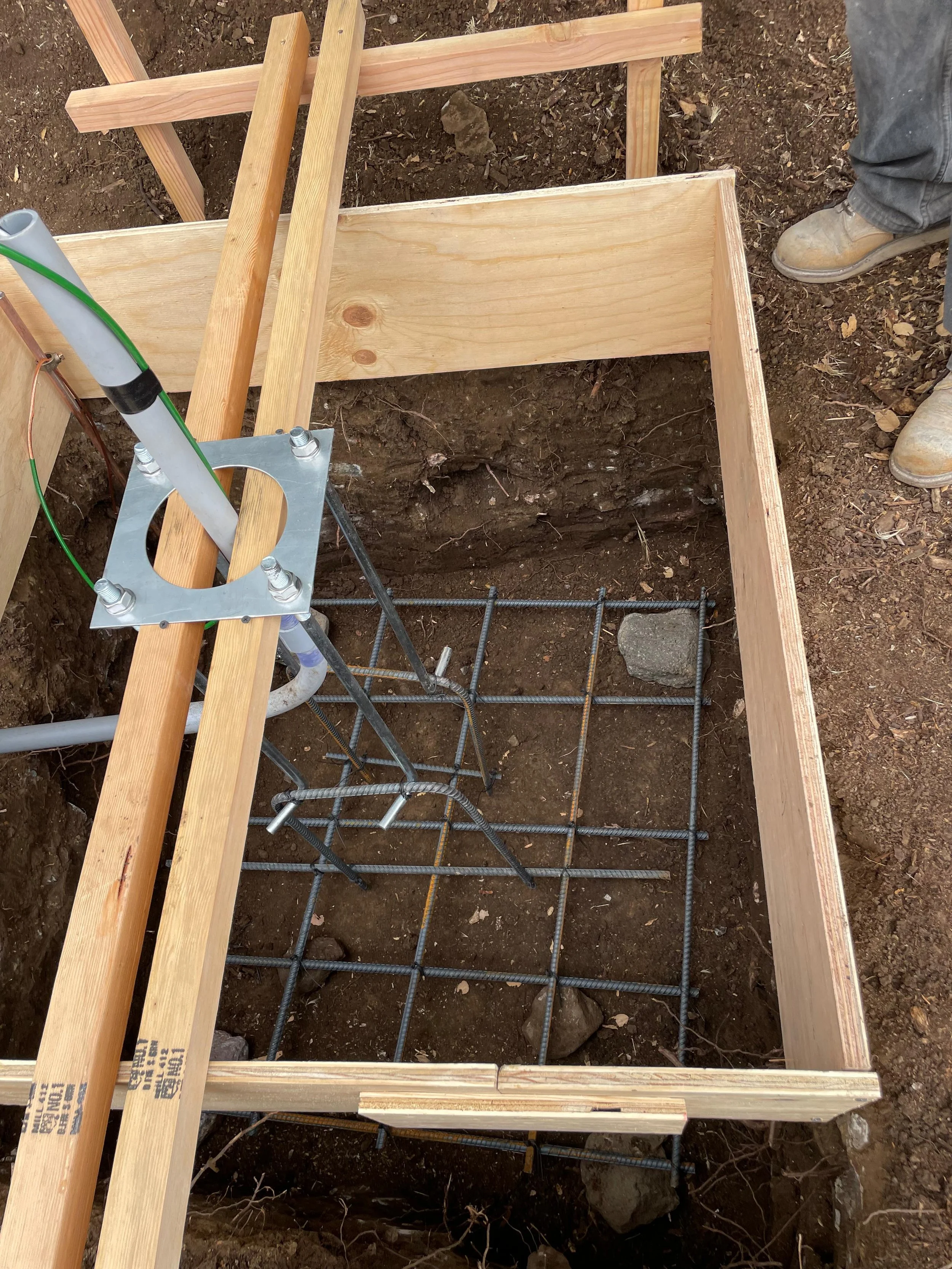 Construction site with wooden framing, rebar, and machinery on dirt ground, indicating early stage of building foundation.