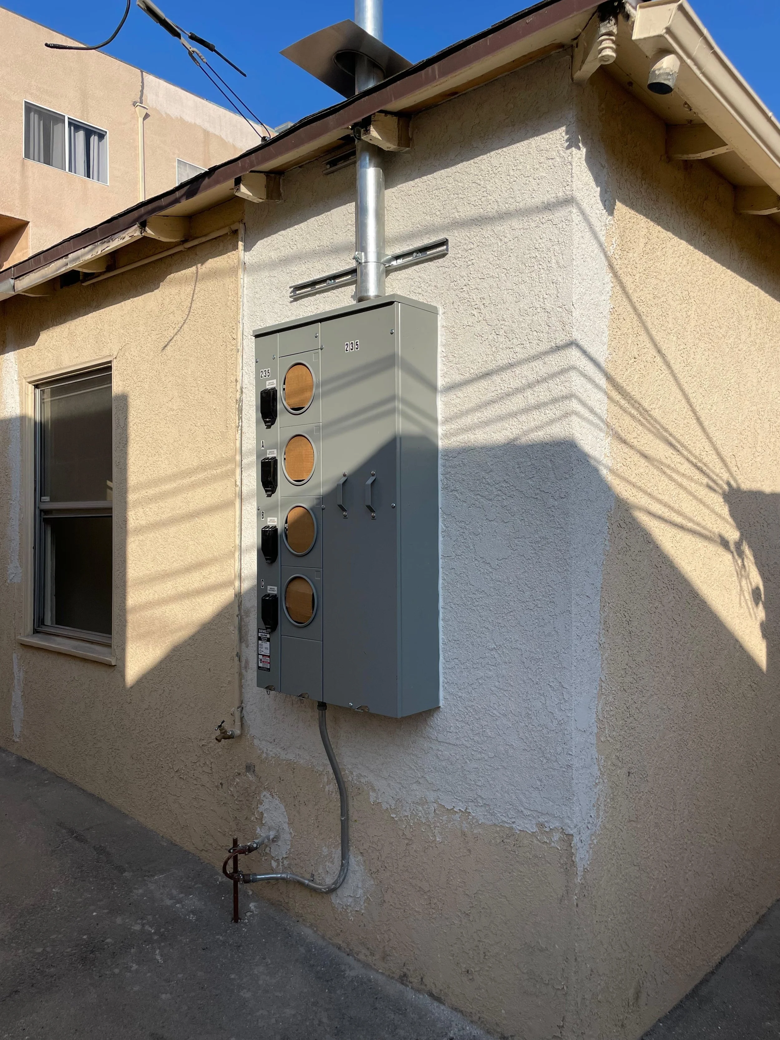 Outdoor electrical meter box mounted on a beige stucco wall of a building, with pipes and wiring visible nearby.