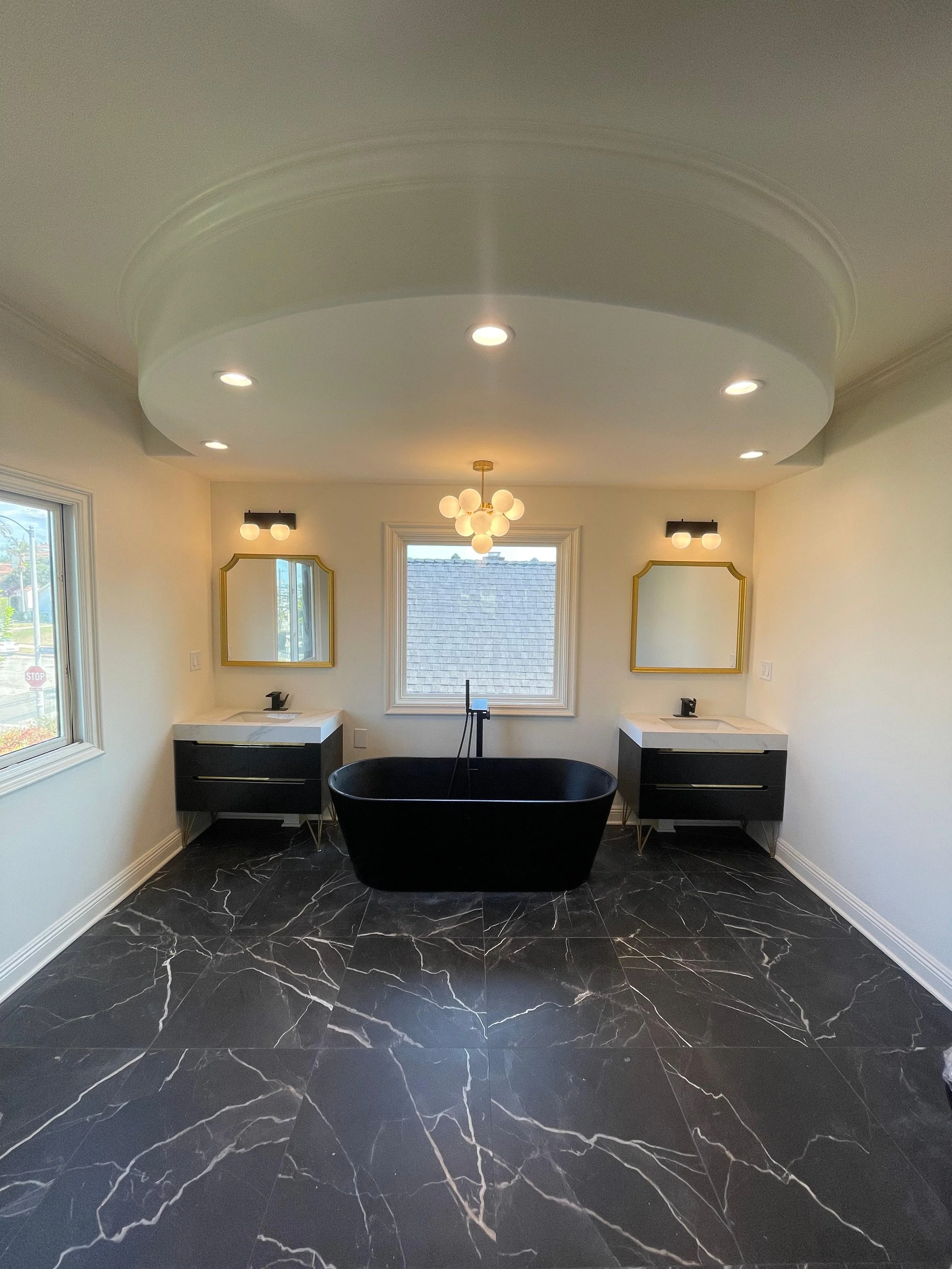 Bathroom with black marble floor, two white vanities with black drawers, gold-framed mirrors, a black freestanding bathtub, and a central window with a view of the roof outside. There is a modern chandelier hanging from the ceiling and recessed light