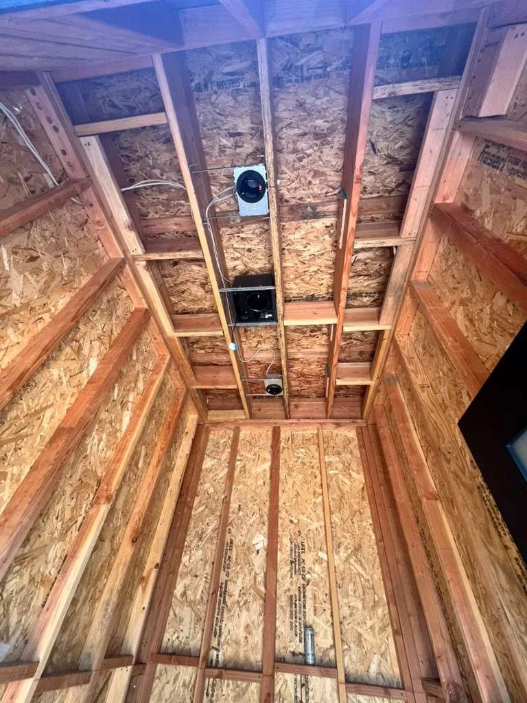 Interior view of a wooden room under construction with exposed OSB walls and ceiling, showing wiring and ventilation ducts.