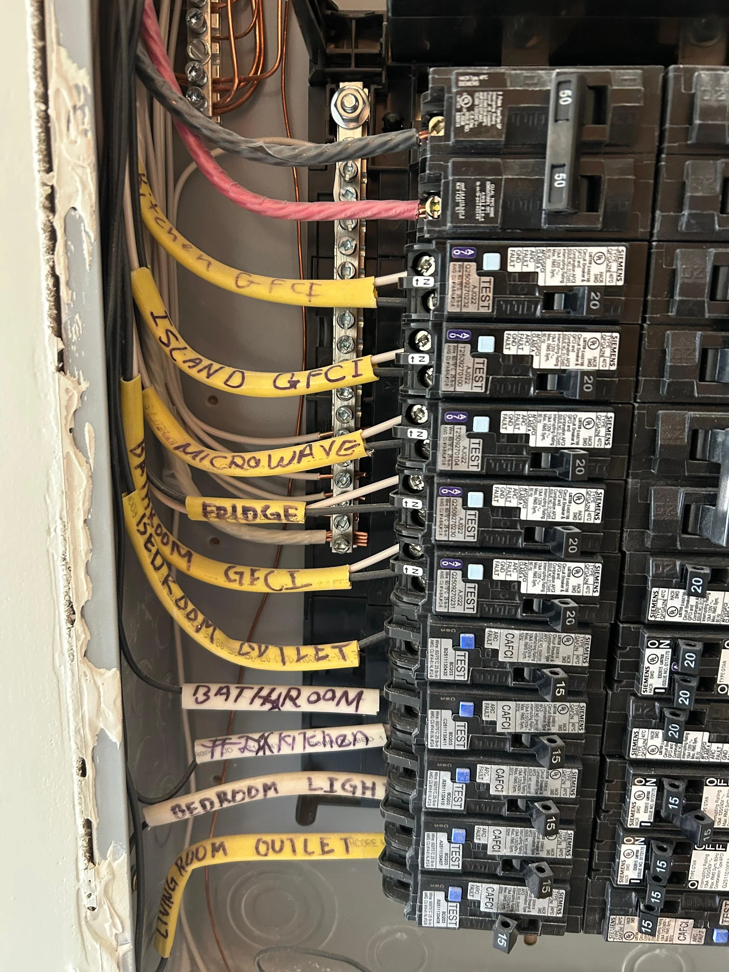 Close-up view of an electrical breaker panel with multiple circuit breakers and color-coded wires labeled with various room and device names such as 'Bathroom', 'Bedroom', 'Outlets', and 'Kitchen'.