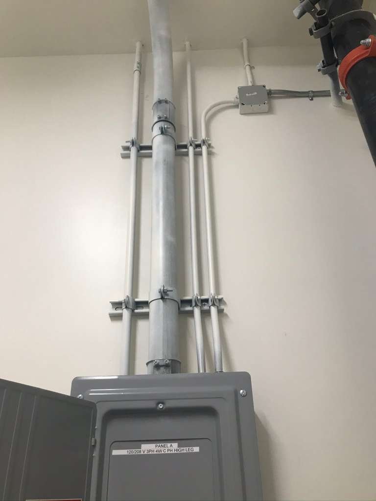 Electrical panel with conduit pipes and a pipe connection on a beige wall in a utility or basement area.