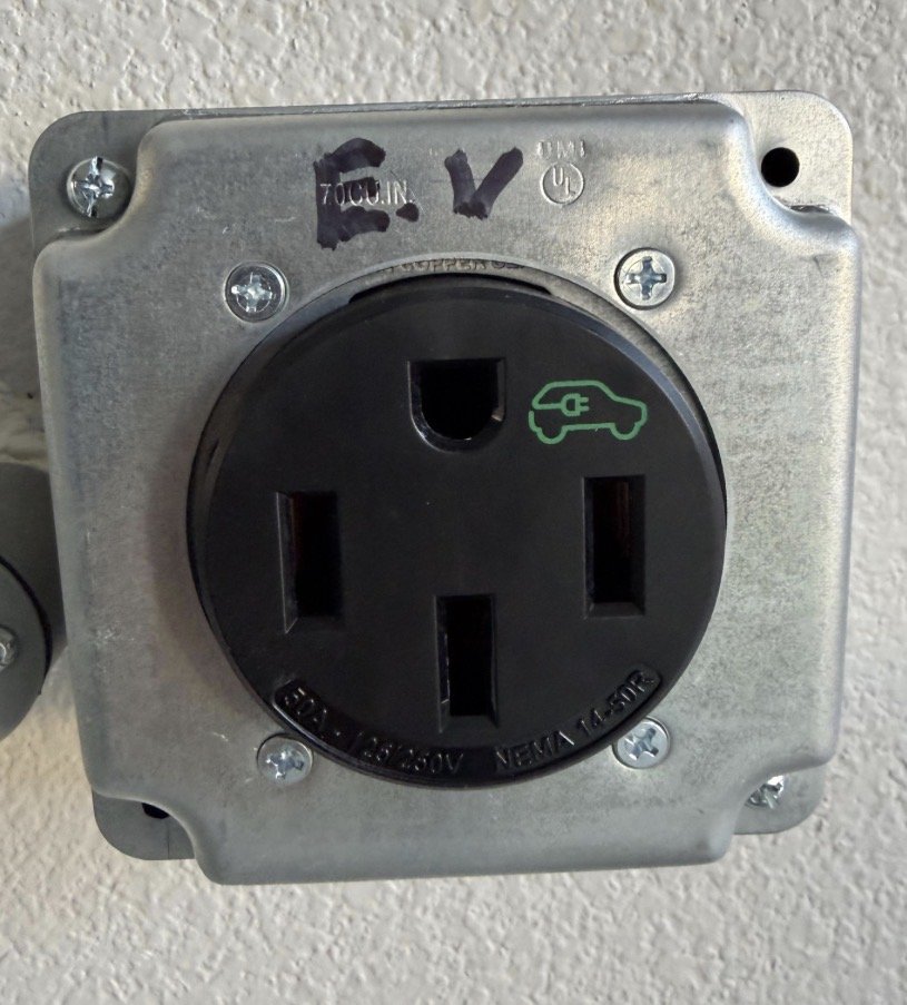 Electrical outlet with an electric vehicle charging symbol on the right side of the socket, mounted on a wall with a metal faceplate and four screws.