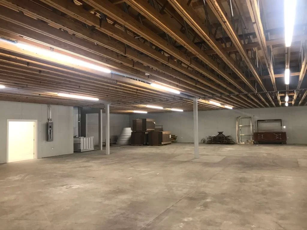 Empty warehouse with unfinished ceiling showing wooden beams and fluorescent lighting, concrete floor, and some stacked boxes and metal shelves in the back.